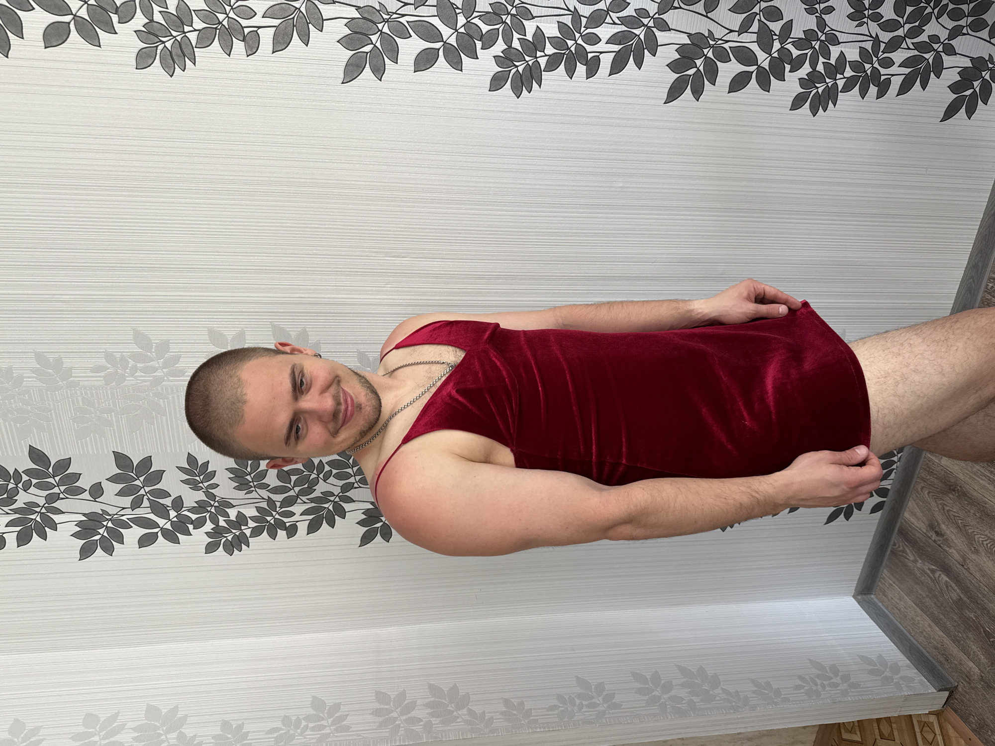 The image depicts a young man, appearing to be in his early twenties, standing indoors in front of a wall with a striped, floral-patterned wallpaper. He has a shaved head and is wearing a red velvet slip dress with thin spaghetti straps and a V-neckline. A silver chain necklace is visible around his neck. His right arm is bent at the elbow, and his hand is resting on the hem of the dress. His left arm is by his side. He is looking towards his left with a slight smirk. The floor is covered with wooden laminate flooring. The scene suggests an informal setting within a residential interior. The location of Sharkan, Russia is not discernible from the visual information provided in the image.