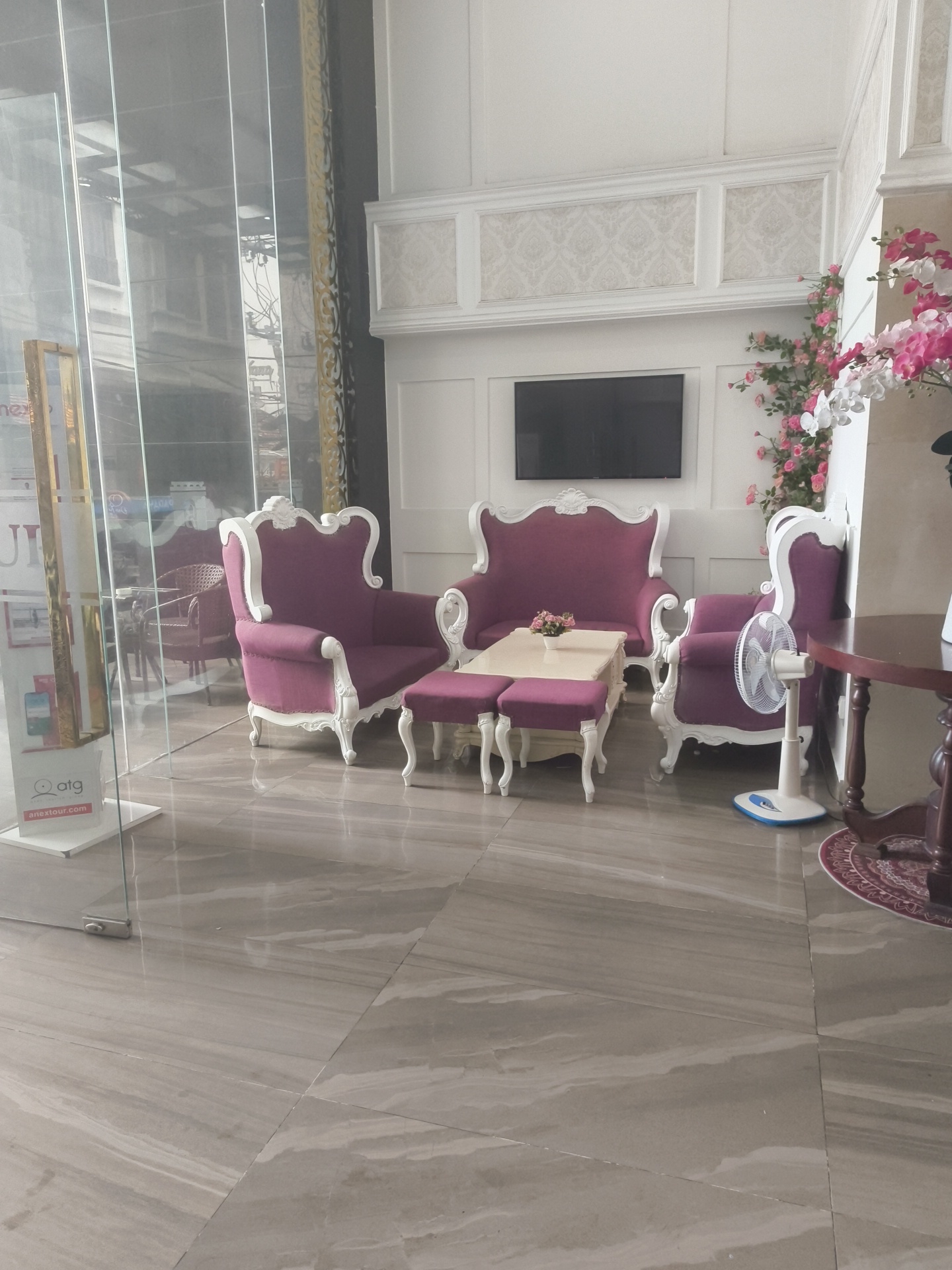 The image shows a hotel lobby in Nha Trang, Vietnam. The lobby features plush purple velvet chairs with ornate white carvings, a small light-colored coffee table with a floral centerpiece, and two matching footstools. A modern flat-screen television is mounted on the wall above a decorative panel. To the right, a white oscillating fan stands near another chair, and a dark wooden table with a circular rug is visible. The floor is tiled with large, gray marble-patterned tiles, and a glass wall with reflections of the exterior is on the left side of the frame. A sign for "anextour.com" is visible near the entrance.