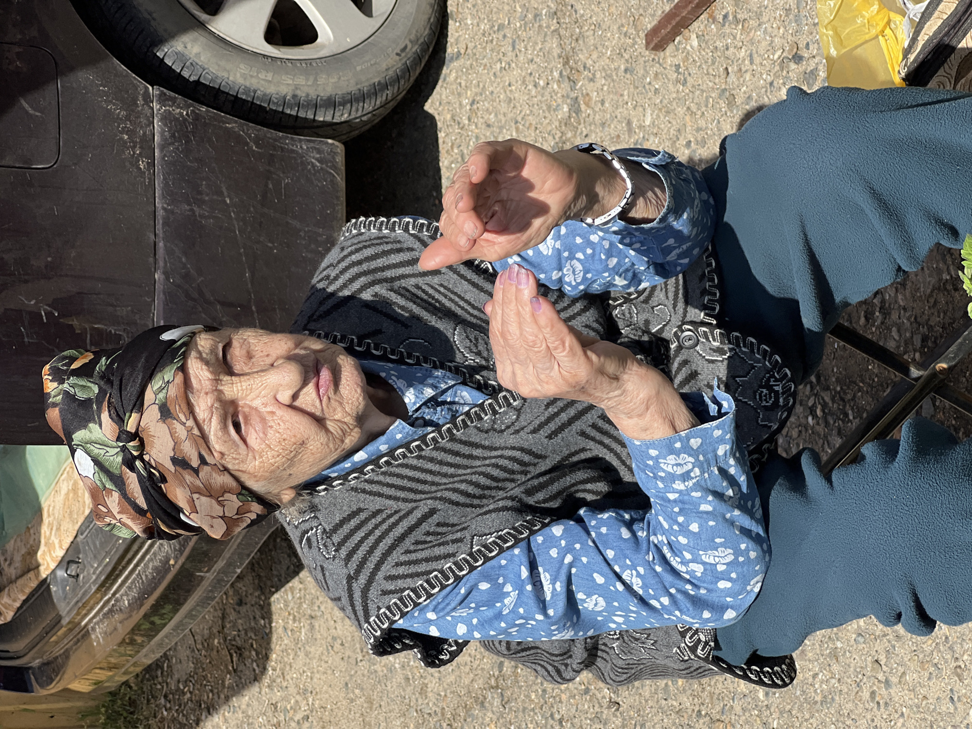 An elderly woman sits outside, partially obscured by a dark-colored vehicle, on a sunny day in what appears to be a street in Goryachiy Klyuch, Russia. She wears a floral headscarf, a blue patterned shirt, and a gray vest with decorative edging. Her hands are clasped in front of her. The surroundings include a paved surface and, in the background, a hint of foliage and other objects.