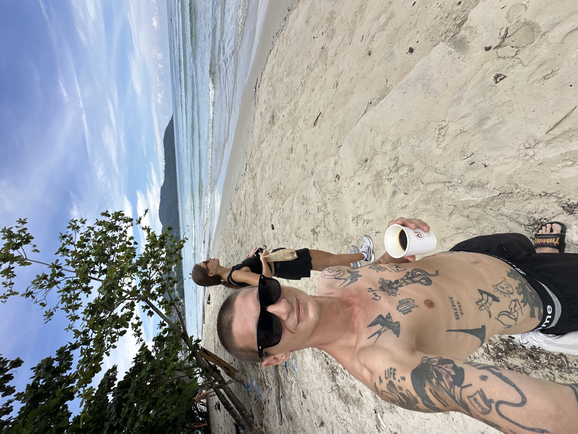 A man with extensive tattoos and sunglasses takes a selfie on a sandy beach in Karon, Thailand, holding a white cup of coffee. He is wearing black shorts with a white waistband. Behind him, a woman in a black tank top and skirt stands looking out at the ocean, holding a light-colored bag. The ocean waves gently lap the shore under a partially cloudy blue sky. Trees with green leaves are visible on the left side of the frame, casting shadows on the sand. The beach appears to have scattered debris.
