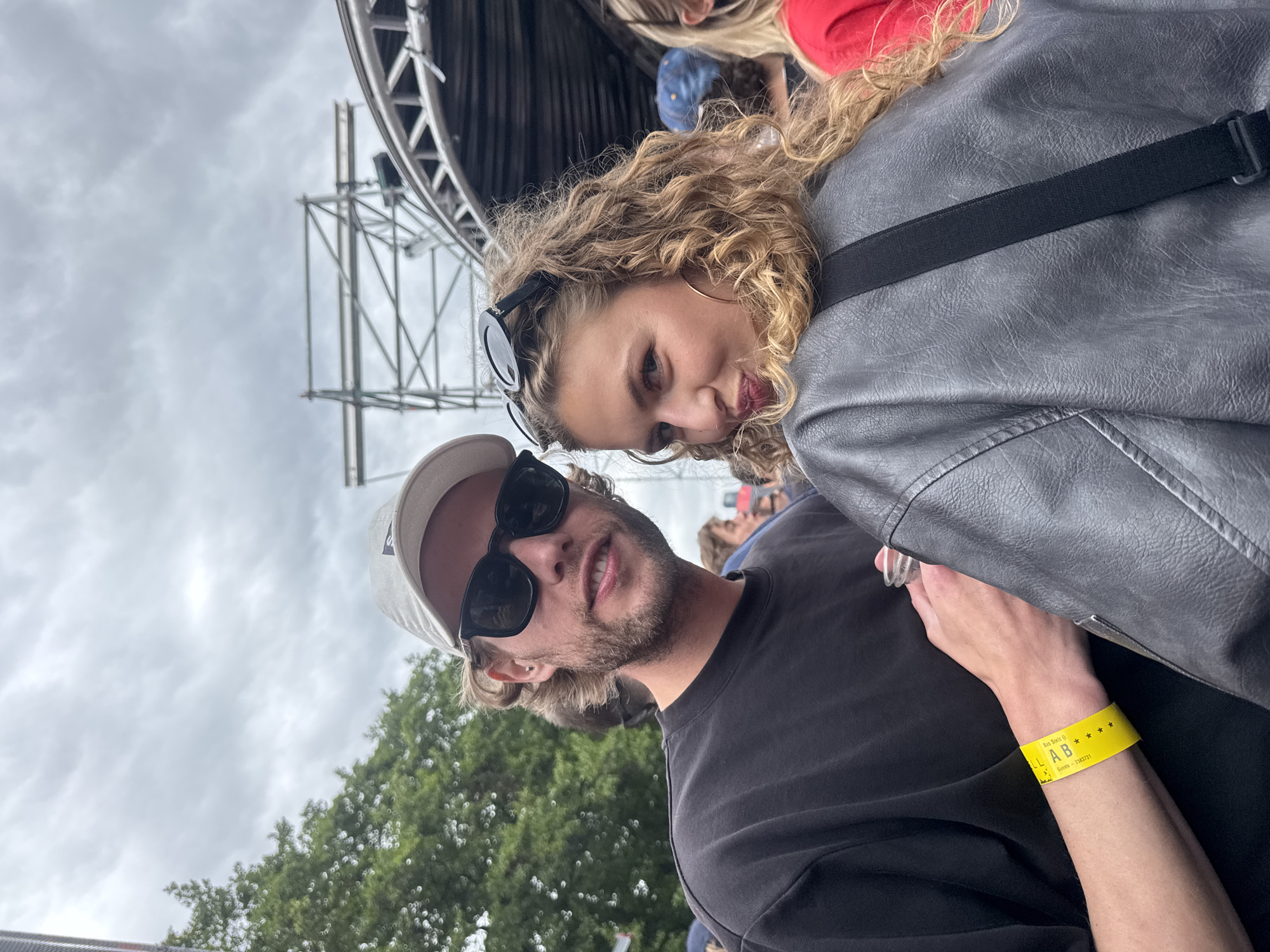 Two individuals, a man and a woman, are posing for a selfie outdoors. The man, on the left, is wearing a black t-shirt, a white baseball cap, and sunglasses. He has light brown hair and a beard, and is looking at the camera. A yellow wristband with black lettering is visible on his left wrist. The woman, on the right, is wearing a grey leather jacket and has blonde, curly hair. She is looking over her shoulder at the camera, with sunglasses perched on her head. Both are outdoors, with a stage structure and a crowd of people visible in the background, suggesting a festival or concert. The sky is overcast. There is no clear indication of the city of Zaandam, Netherlands in the image itself.