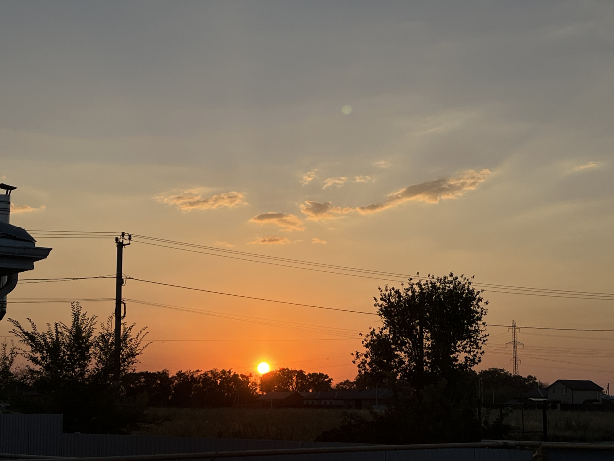 The image depicts a rural landscape at sunset, likely within the Taganrog City District of Russia. The sky transitions from a pale gray at the top to a vibrant orange near the horizon, dotted with a few wispy clouds illuminated by the setting sun. A bright, circular sun is visible low in the sky, casting a warm glow.
In the foreground, a dark silhouette of trees and foliage frames the scene. A metallic fence runs along the bottom of the image. To the left, the corner of a building with a white roof and a chimney is visible. A utility pole with electrical wires stretches across the mid-ground, intersected by several power lines. To the right, a silhouette of a larger tree stands prominently, and further in the distance, a tall electrical pylon and a small house are discernible against the twilight sky. The overall impression is one of a peaceful, rural evening.