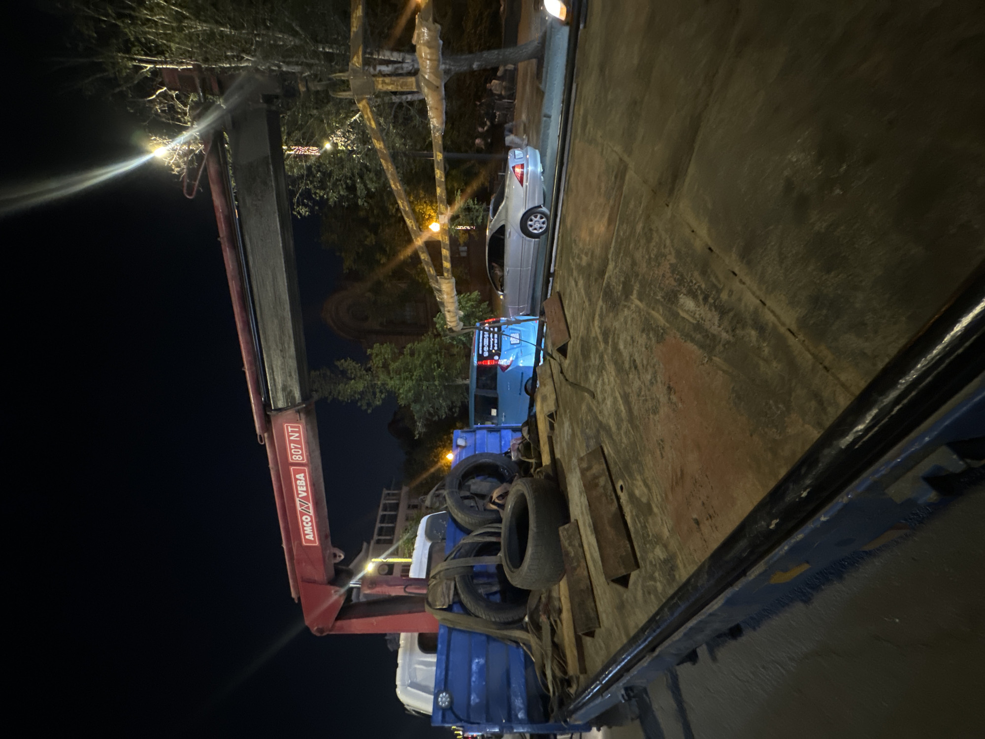 This image depicts a flatbed tow truck at night, with its crane extended. The truck is loaded with several tires and straps. In the background, a light blue minivan and a silver sedan are visible. The scene appears to be in an urban setting, with trees and buildings present. The tow truck has the branding "AMCO VEBA 807 NT" on its crane.
There is no information in the image that would allow for the specific identification of the location as Krasnoyarsk, Russia.