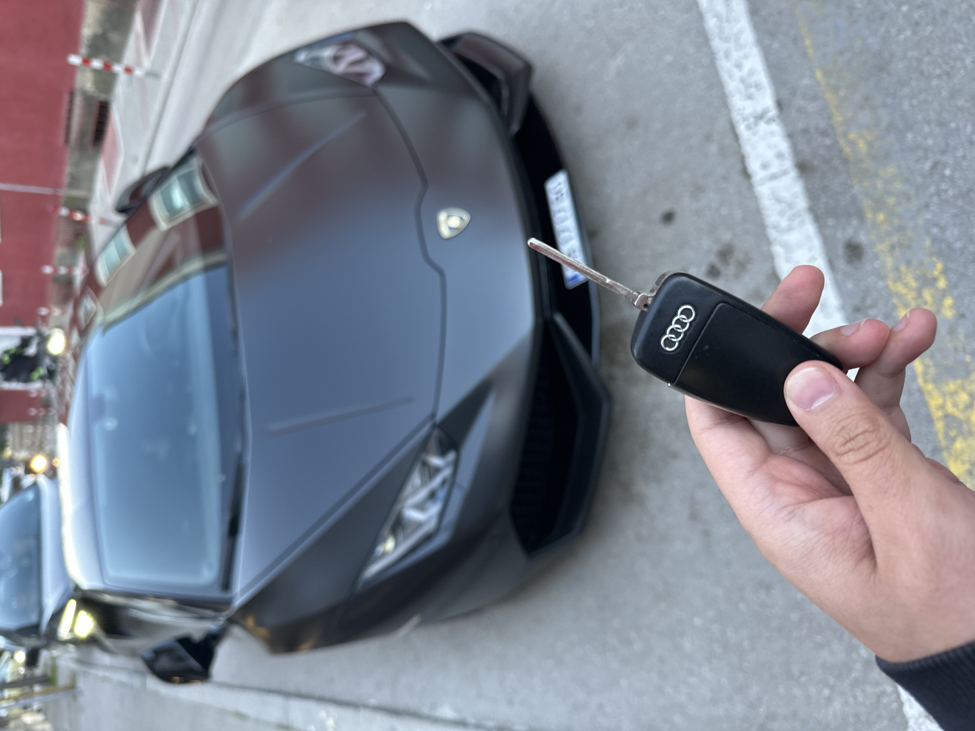 A matte black Lamborghini sports car is parked on a gray concrete surface, likely a street or parking area. An Audi car key is held in a person's right hand near the Lamborghini. The scene appears to be set outdoors, with a building visible in the background. There's no immediate confirmation of the location as Veliko Tarnovo, Bulgaria.