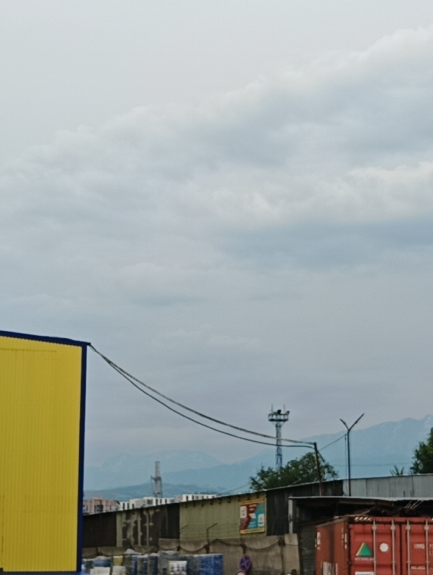 This is a landscape shot taken outdoors in a semi-urban or industrial area with a mountain range in the background. The sky is overcast with thick, grey clouds, suggesting it might be early morning, late afternoon, or simply a cloudy day. The overall mood is somewhat muted due to the weather.
In the foreground, a bright yellow corrugated metal structure, likely a building or container, dominates the left side of the frame. Its blue trim adds a contrasting element. Two dark wires stretch diagonally across the middle of the image, connecting to the yellow structure and extending out of frame.
Behind the yellow structure, various low-lying buildings with metal roofs and walls are visible. Some appear aged and weathered, with a mix of dark and lighter colors. In front of these buildings, stacks of what appear to be pallets or crates are arranged, some covered with blue tarpaulin.
Further in the background, a tall, slender radio tower or communication mast with a small platform at the top stands against the hazy mountain backdrop. A streetlamp with a distinctive Y-shaped design is also visible to the right of the tower. The mountain range itself is softened by atmospheric haze, with a lighter blue hue.
There is a sign with colorful graphics on one of the buildings, but the text is illegible. A red shipping container is partially visible on the far right, with some green and white lettering on its side, also too indistinct to read. No people are clearly visible in the frame. The scene appears to be a functional area, possibly a storage yard, warehouse district, or a light industrial zone.