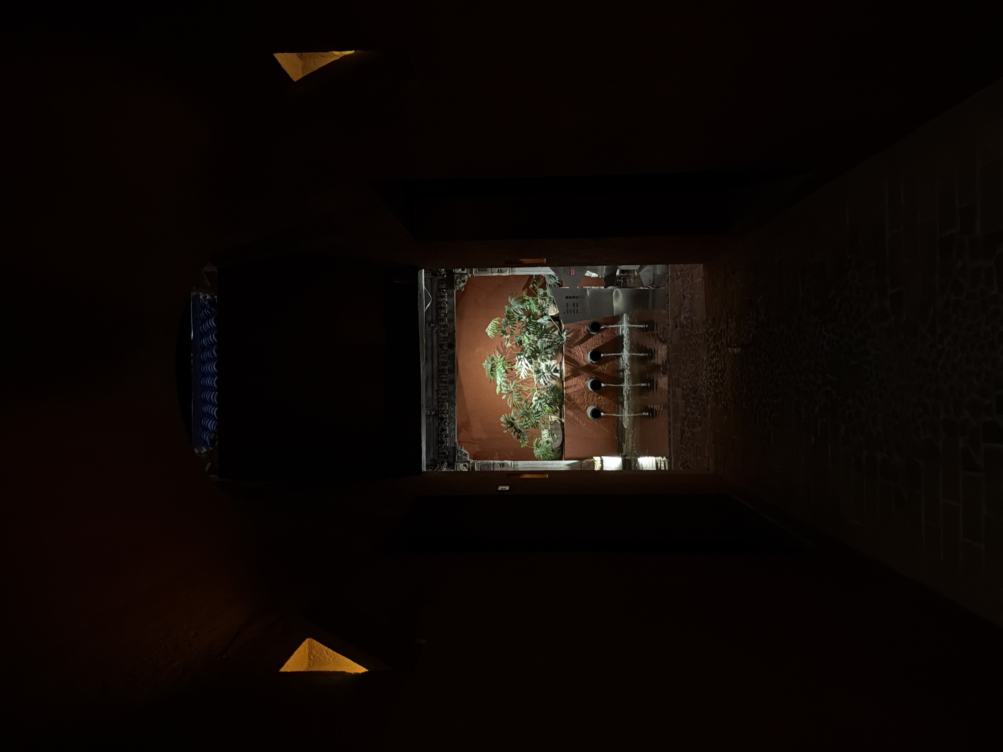 A narrow, dark alleyway in Puebla, Mexico, with brick or stone walls leading to a brightly lit courtyard. The courtyard features a reddish-brown wall adorned with greenery and what appears to be a water feature with several vertical pipes. At the other end of the alleyway, a corner shows architectural detail, possibly a window or decorative element. There are two triangular lights illuminating the walls of the alleyway in the foreground.