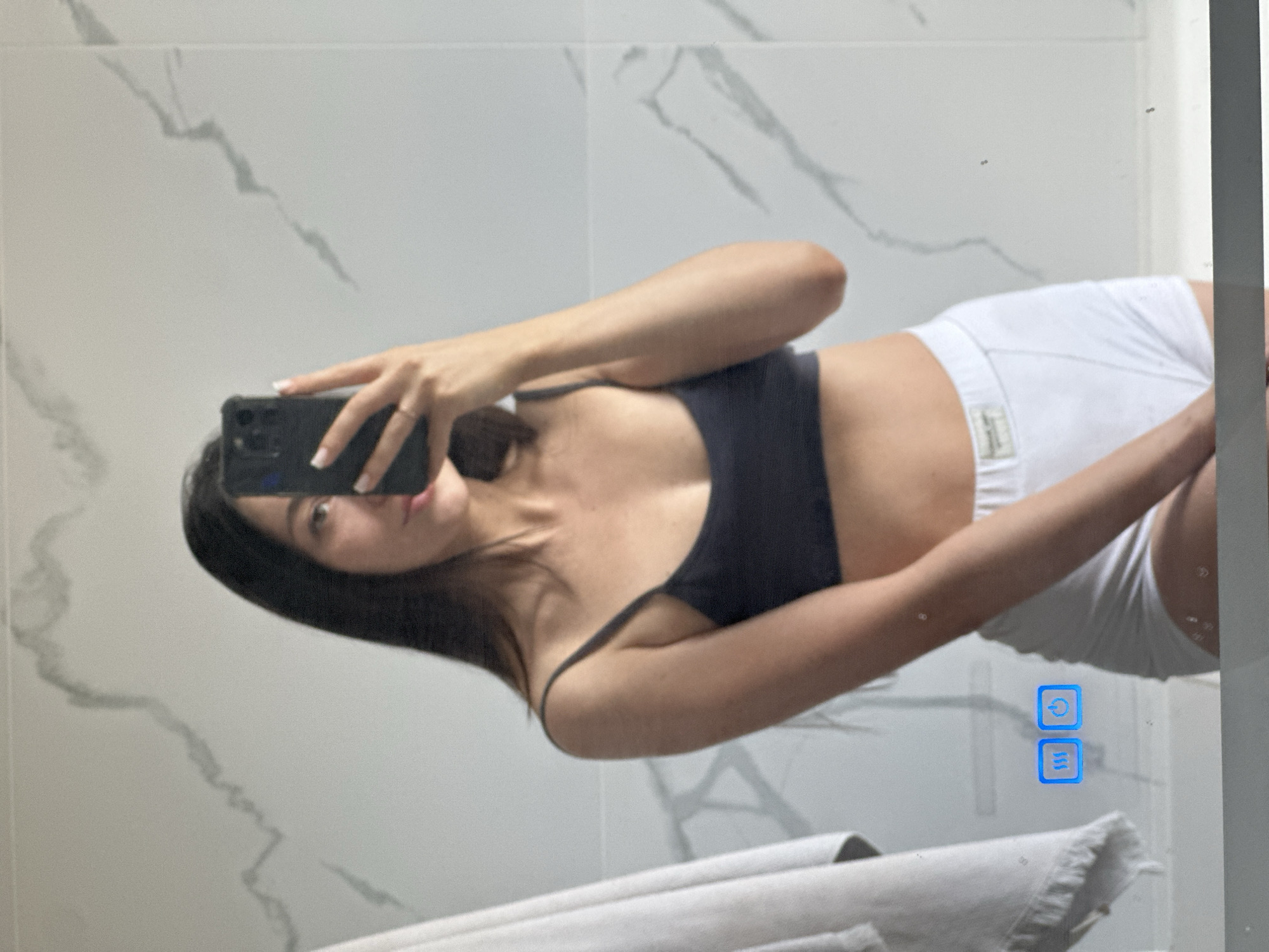 A young woman with long dark hair takes a mirror selfie in a bathroom. She is wearing a dark sports bra and white shorts. Her left arm is raised, holding a black smartphone to capture the image. Her right hand is resting on her hip. In the background, a white towel hangs on the left side of the frame, and a marble-patterned wall is visible behind her. Two illuminated blue icons, one resembling waves and the other a power symbol, are integrated into the mirror. The scene appears to be in a modern bathroom, but no specific location in Эстачи, Russia, can be determined from the image alone.