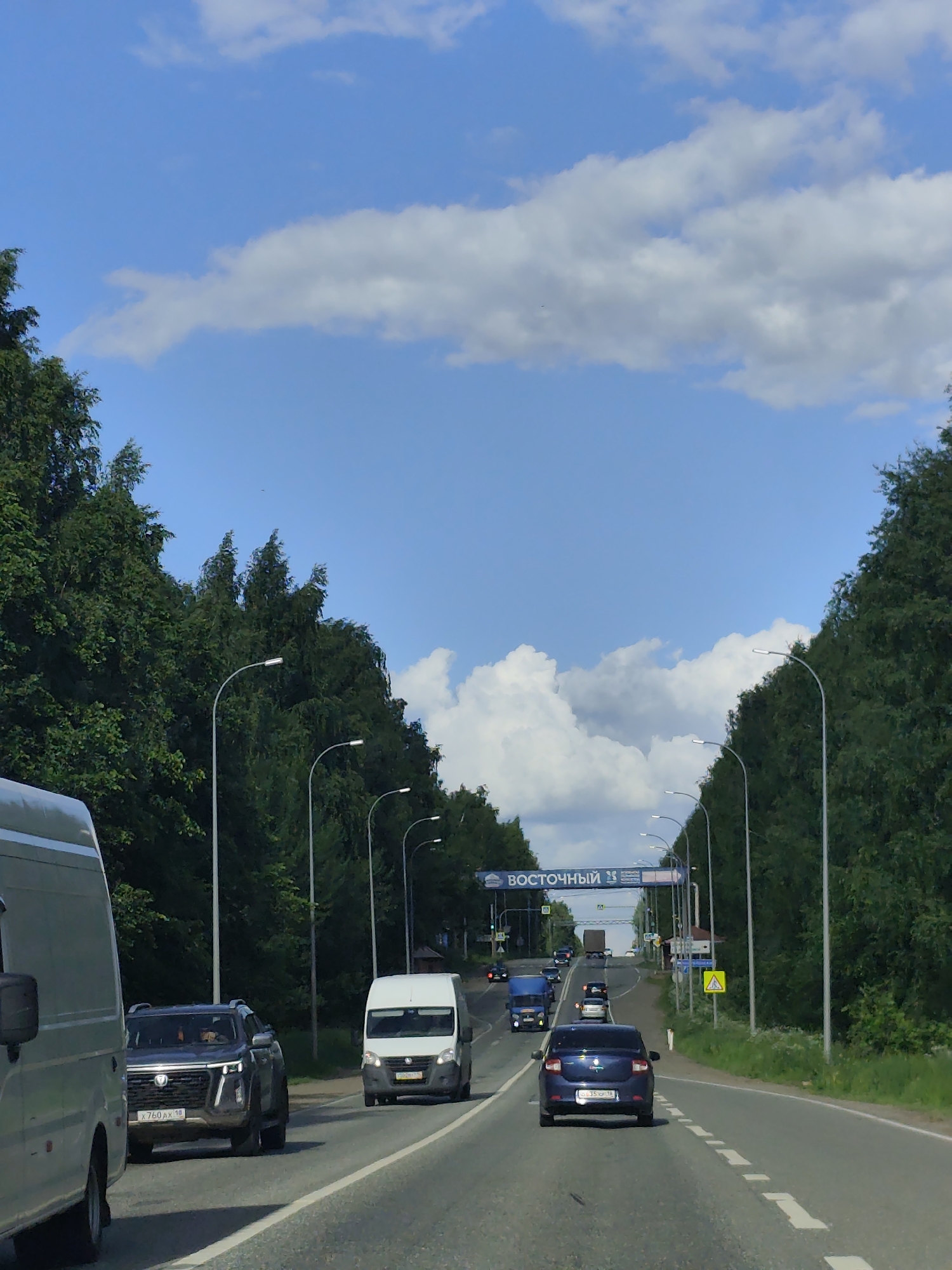 The image depicts a sunny day on a highway, likely near the city of Italmas, Russia, given the context. The highway is lined with dense green trees on both sides. A blue sign arching over the road reads "ВОСТОЧНЫЙ" followed by a number, possibly indicating a district or landmark. Several vehicles are visible on the road, including a white van, a dark SUV with license plate "X 760 AX 18", a blue sedan with license plate "X 351 HP 18", and other cars and trucks further down the road. Streetlights are positioned along the side of the highway. The sky is bright blue with scattered white clouds. The overall scene suggests a typical day of travel on a Russian highway.