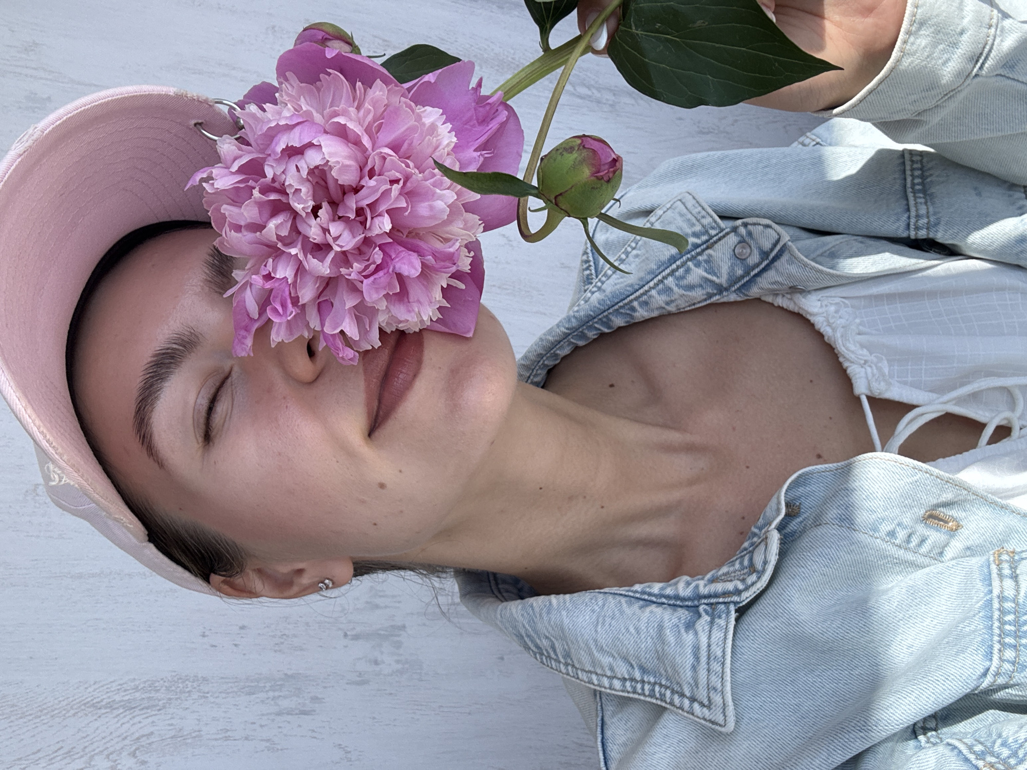 In this close-up, medium-shot photograph, a young woman is depicted against a light-colored, textured background, possibly a wall. She is wearing a light blue denim jacket over a white top with lace trim. On her head, she has a pale pink baseball cap. Her eyes are closed, and she is holding a large, vibrant pink peony flower in front of her face, obscuring her nose and mouth. The flower's petals are lush and layered, with a lighter pink hue towards the center. A single green bud is visible on the stem of the peony, along with a few green leaves. The woman's skin appears smooth, with a hint of blush on her cheeks and her lips painted a soft, rosy color. She is wearing a small, silver hoop earring in her visible ear. The lighting suggests a bright, possibly outdoor, environment, casting subtle shadows on her neck and shoulder. The image focuses on the woman's upper body and the striking contrast between her skin, the denim, and the delicate beauty of the peony.
