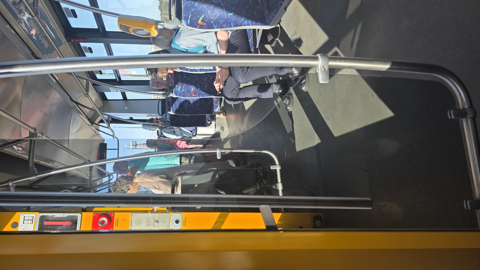 The image depicts the interior of a public transport vehicle, likely a tram or bus, with passengers seated and standing. The overall lighting suggests it is daytime, with sunlight casting shadows across the floor and seats. The vehicle's interior features yellow and silver metallic accents, a common design element. Several individuals are visible, some facing forward, others looking at their phones. The seats are upholstered in a patterned fabric. On the left side of the image, a yellow panel contains a red emergency stop button and other controls. The scene appears to be within the city of Riga, Latvia, given the visible signage and architectural style.