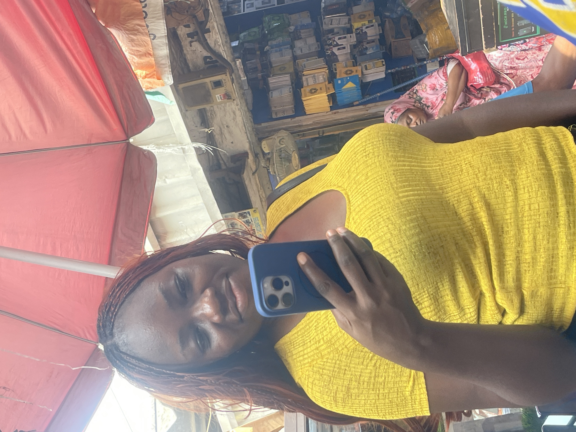 An image captures a scene within what appears to be a bustling market in Abuja, Nigeria, during the daytime. The primary subject is a woman in the foreground, taking a selfie. She has dark skin, long reddish-brown braided hair, and is wearing a bright yellow, ribbed, off-the-shoulder top. She holds a blue smartphone with a prominent camera array, angled towards herself with a subtle smile.
In the background, a large red umbrella or canopy provides shade or cover. Behind the selfie-taker, the market stall is densely packed with various goods. Shelves are laden with numerous boxes and packaged items, many appearing to be electronic accessories or small general merchandise. Some boxes prominently display the word "Gled" or "Glad," while a larger black box shows "KICH PRO." A white industrial fan is visible among the goods, suggesting the need for air circulation.
Further into the background, another woman is visible, seemingly resting or sleeping. She is covered in a pink floral-patterned garment, possibly a hijab or full-body wrap, with a red bag or item near her head. The overall setting indicates an active commercial environment, characterized by the variety of goods and the informal structure of the stalls, consistent with a local market in West Africa. The bright lighting suggests it is daytime.