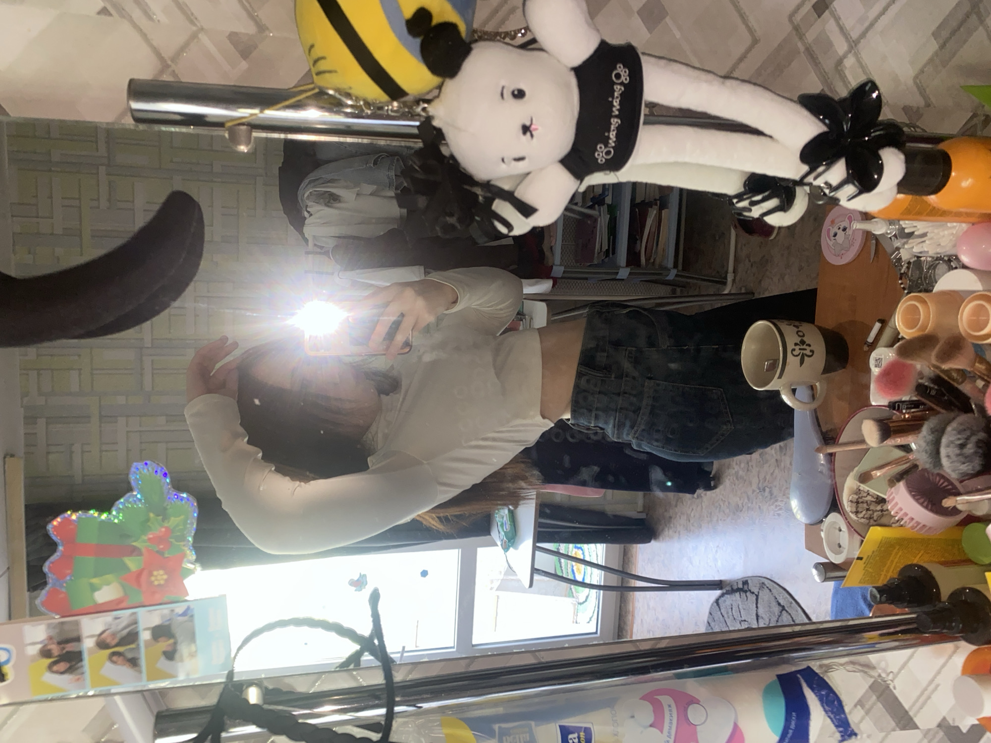 A mirror reflection shows a person taking a selfie. The person is wearing a white long-sleeved shirt and dark jeans. In the background, a window provides natural light. Various objects are visible around the mirror, including a stuffed animal, a mug, makeup brushes, and hair products. The location is Unknown, Unknown.