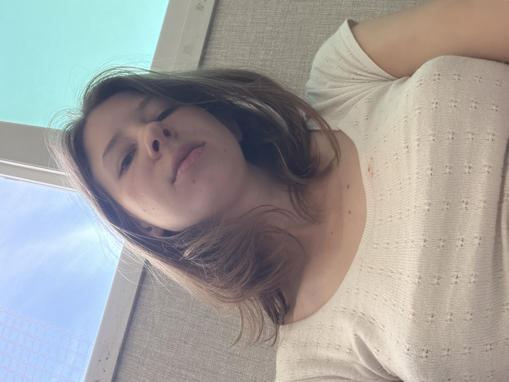 A young woman with shoulder-length brown hair and fair skin is pictured from the chest up, looking down at the camera. She is wearing a white, short-sleeved, textured shirt. A small red mark is visible on the shirt, near her left collarbone. She has a mole on her left cheek and another on her upper lip. The background consists of a textured wall, a window with a blue sky visible through it, and a white frame. The scene appears to be indoors, with natural light coming from the window.