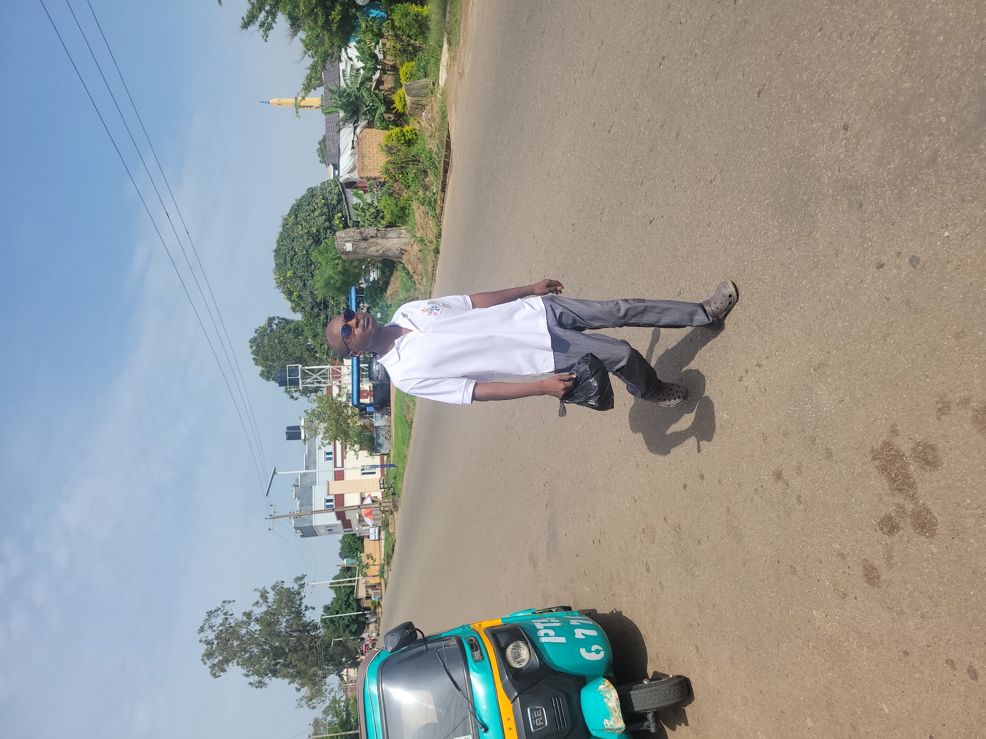 A man is walking across a paved road in Jos, Nigeria, carrying a black bag. He is wearing a white polo shirt and grey trousers. To his left, a turquoise auto-rickshaw is parked on the side of the road. The background shows buildings, trees, and power lines under a clear blue sky.