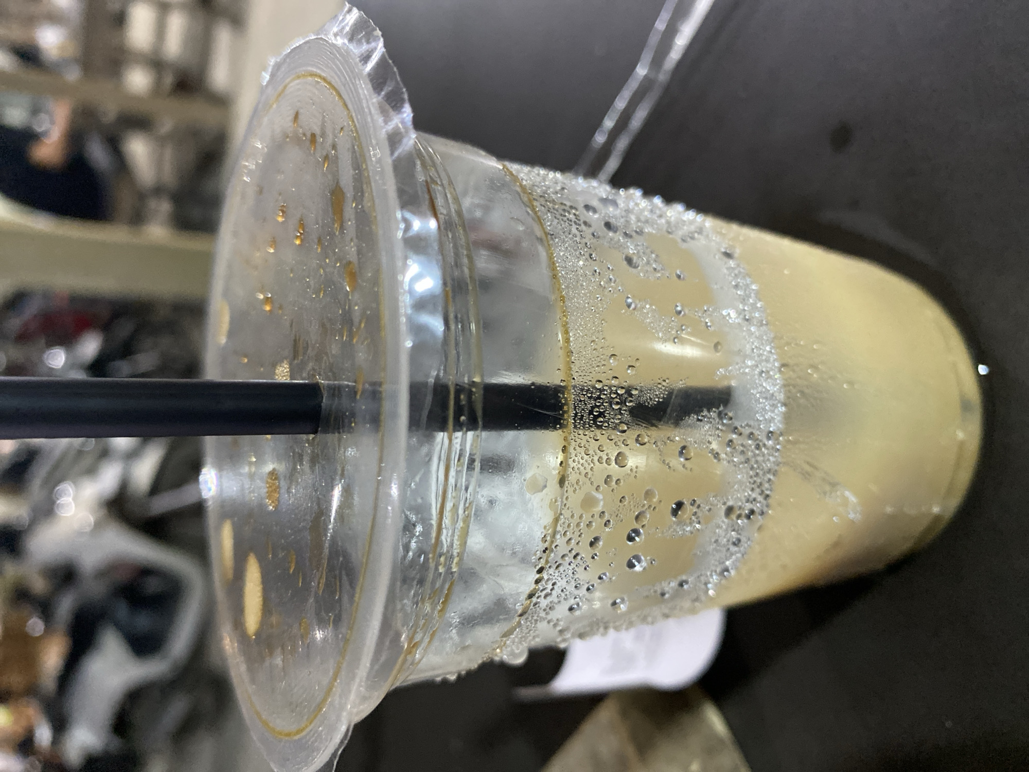 The media file presents a close-up view of a clear plastic cup containing a light-colored, possibly milky, beverage. The image is rotated 90 degrees clockwise, making the cup appear to be resting on its side on a dark, reflective surface. A black straw is inserted through the clear plastic lid into the drink.
A prominent feature is the heavy condensation on both the exterior and interior of the cup, manifested as numerous small water droplets, indicating the beverage is cold. Some darker, possibly brown, liquid residue or drips are visible on the underside of the plastic lid, near where it seals the cup. The beverage itself appears to be a uniform light beige or yellow hue.
The background is out of focus, showing blurred shapes and highlights, suggesting an indoor setting like a cafe, restaurant, or perhaps a casual home environment. No specific objects or details in the background offer further clues about the location within Gampongaceh, Indonesia, or the time of day. No text is visible on the cup, straw, or in the surrounding area. The image primarily focuses on the textured details of the cold drink container.