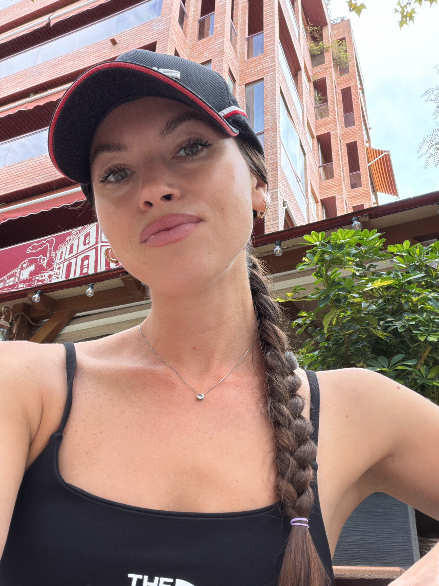 An outdoor selfie shows a woman wearing a black baseball cap and a black tank top. She has a single braid of brown hair extending down her right side. A thin silver necklace with a small pendant is visible around her neck. Her left shoulder and arm are visible. In the background, a multi-story brick building with balconies and awnings is visible. Green foliage is also present in the background, along with exposed light bulbs hanging from a structure. The scene appears to be in Salou, Spain.