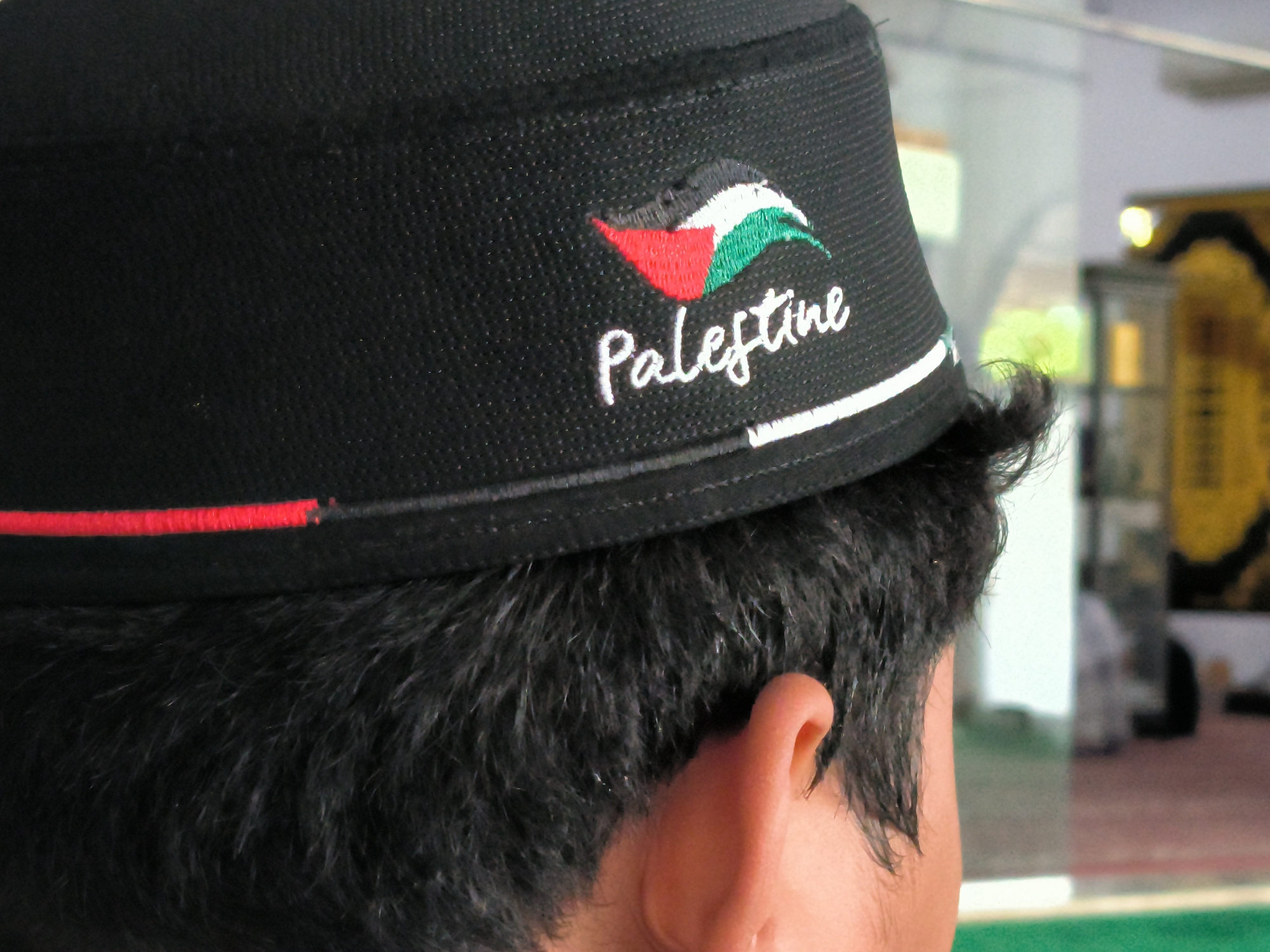The image shows the back of a person's head wearing a black Peci hat. The hat features the colors of the Palestinian flag, along with the word "Palestine" embroidered in white. The person has dark hair and an ear is visible. The background appears to be an interior space, possibly a mosque or religious building. The location is Lhokseumawe, Indonesia.