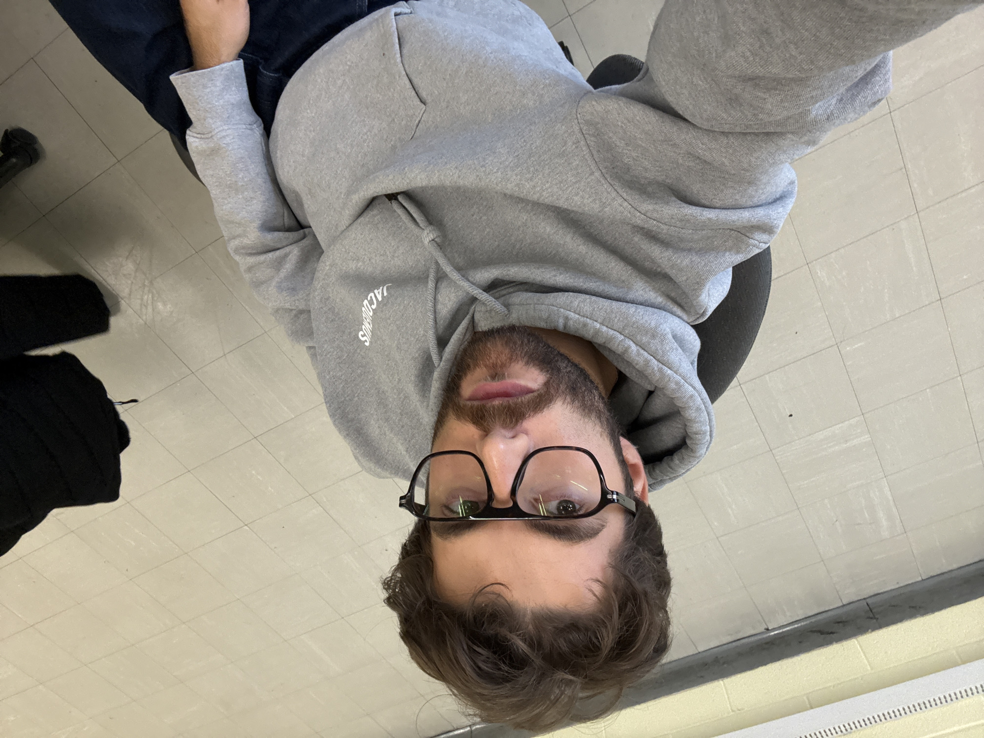 The image is a selfie taken from a high angle, showing a person wearing glasses and a grey hooded sweatshirt with the word "JACQUEMUS" printed on it. The person has a beard and is looking directly at the camera. The background consists of a light-colored tiled floor with a black object to the left of the person. There is no explicit evidence to indicate the city of Montreal. The environment appears to be indoors.