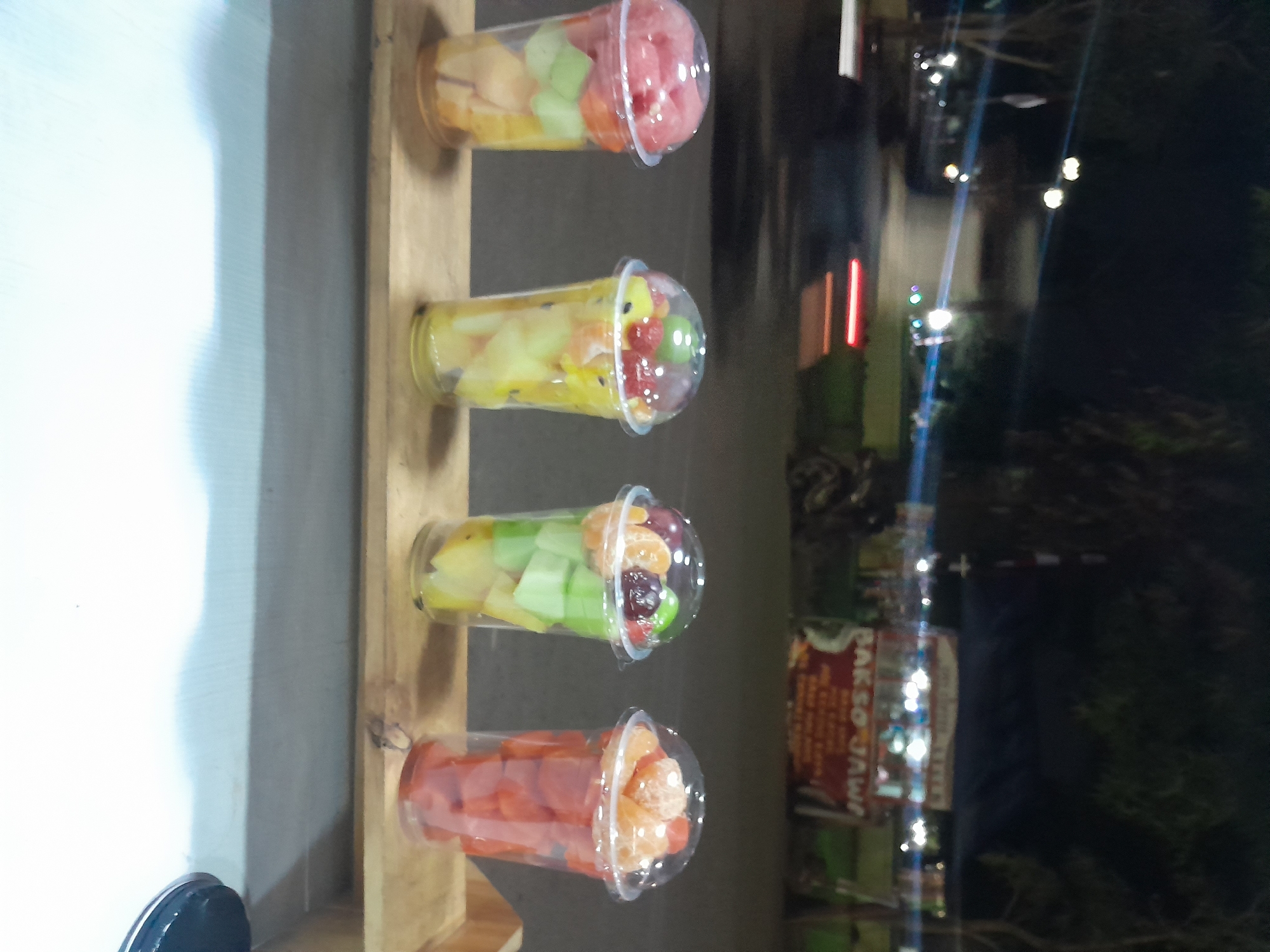 Four clear plastic cups filled with diced fruit are displayed on a wooden plank. The cups are arranged from left to right, with each cup containing a different assortment of fruits. The first cup has red and pink fruit, possibly watermelon, at the top, and lighter colored fruit below. The second cup contains yellow, green, and red fruit. The third cup has green and orange pieces, with some dark purple grapes and a green sphere on top. The fourth cup is filled with orange-colored fruit cubes, appearing to be papaya or cantaloupe. In the background, there is a food stall with red signage that reads "BAKSO JAWA". The scene appears to be outdoors at night, with artificial lights illuminating the area. There are blurred shapes in the background that suggest vehicles and trees. The location is identified as Slawi, Indonesia.