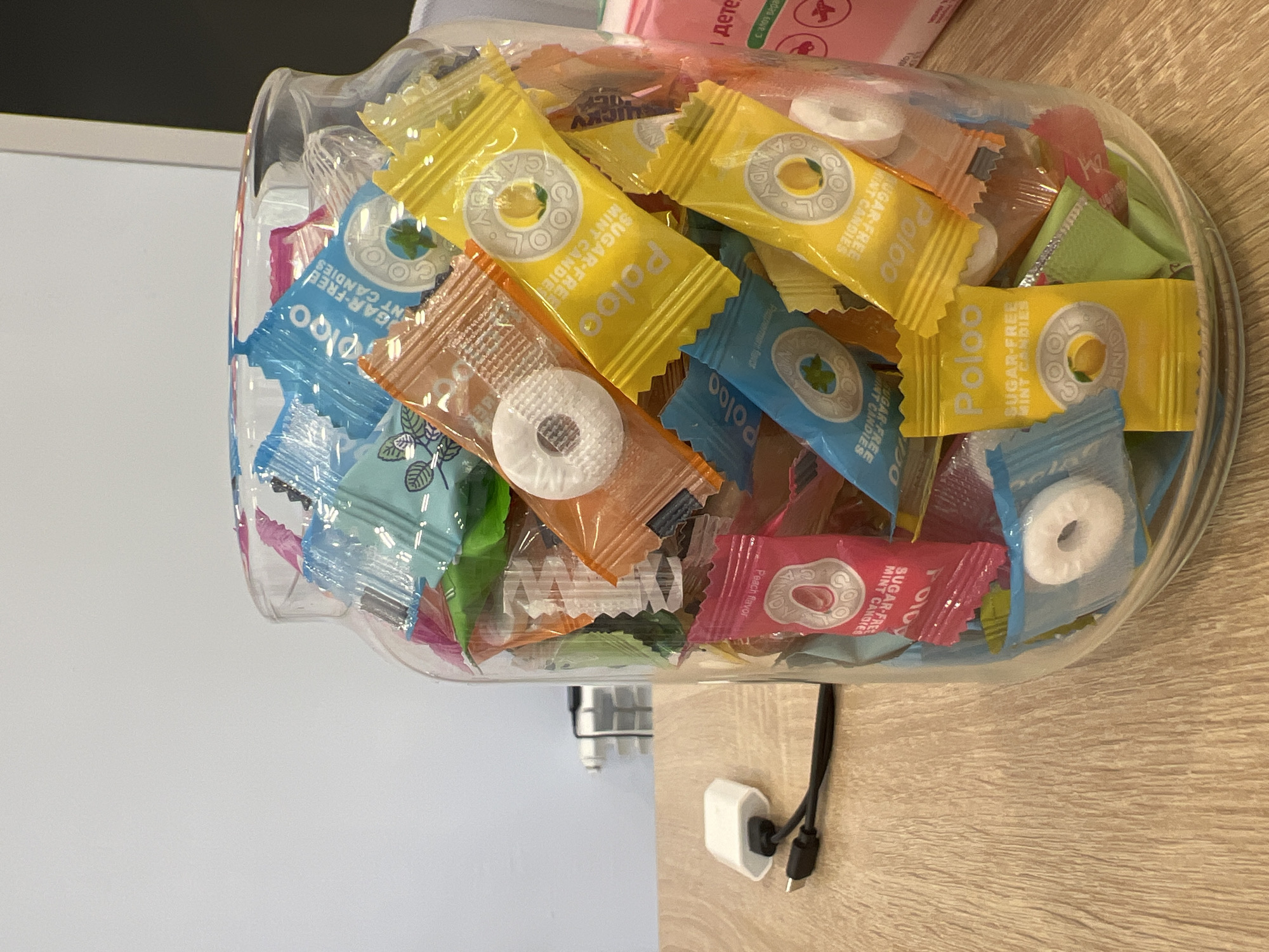 The image shows a glass jar filled with individually wrapped candies. The candies are of various colors, including blue, yellow, orange, and pink, and many have the "Polo" logo visible. Some of the candies appear to be mint flavored. A white USB adapter with a black cable is connected and lying on a wooden surface in the foreground to the left of the jar. The background shows a white wall and a radiator. The scene is indoors, on a wooden table. There is no indication of the city of Bataysk, Russia, or any specific location.