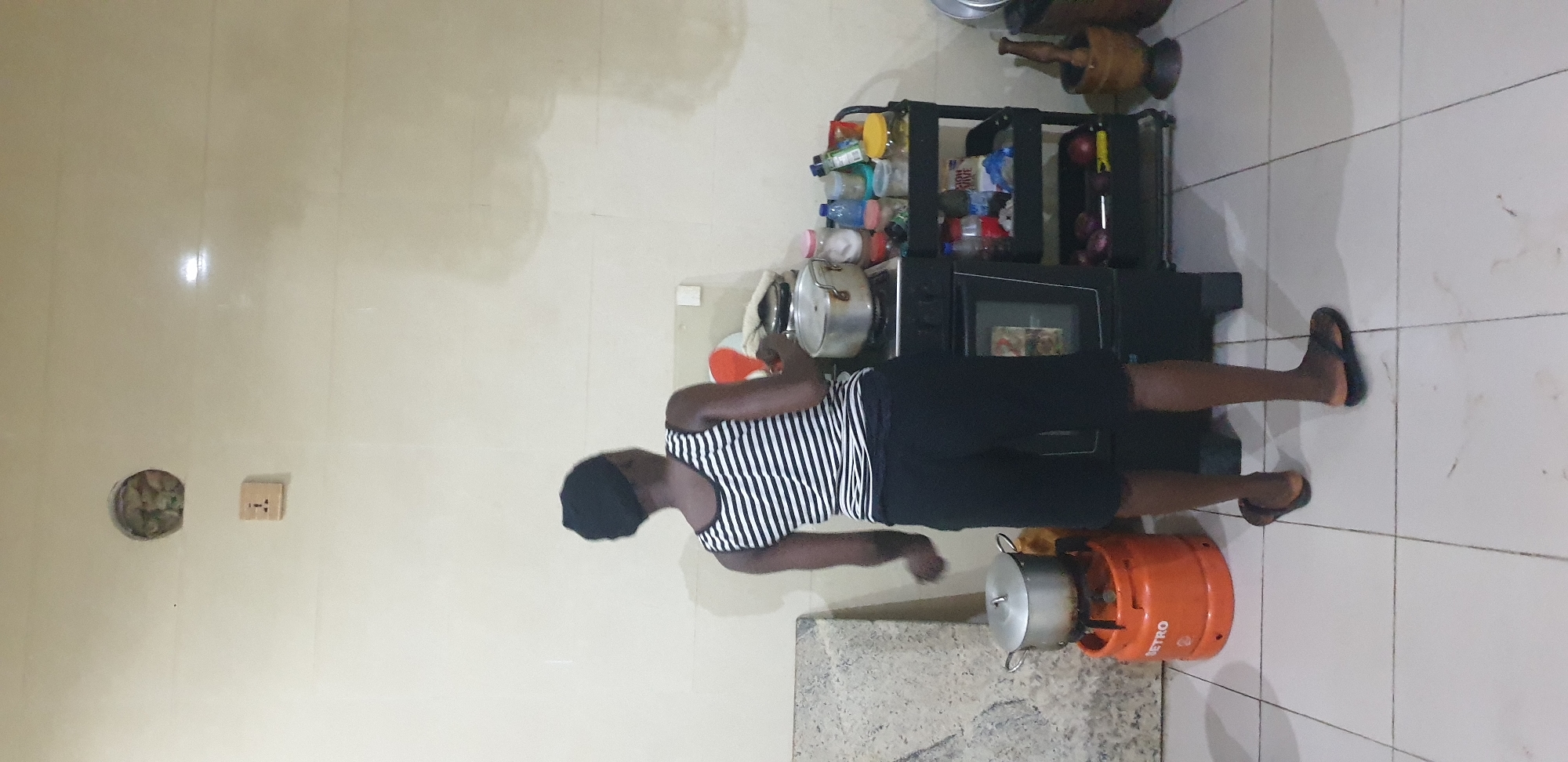 The image depicts an indoor kitchen scene in Unknown, Unknown, featuring an individual facing away from the camera, engaged in cooking. A person with dark skin, wearing a black and white striped tank top, black shorts, and black flip-flops, stands in the center-left, with their back mostly to the viewer. They are positioned in front of a black free-standing stove with an oven, upon which a silver pot with a lid rests. To the left of the stove, an orange "METRO" brand gas cylinder supports a single burner with another silver pot on top. To the right of the stove, a black, three-tiered rolling cart holds various food containers, bottles, and produce, including red onions on the bottom shelf. On the far right, a wooden mortar and pestle are visible. The walls are covered in light-colored tiles, featuring an electrical outlet with three pins above and to the left of the stove, and an irregular circular hole higher up on the wall revealing internal material. A light gray, rectangular slab leans against the wall behind the gas cylinder. The floor is covered in light-colored tiles with visible grout lines. The room is illuminated by artificial light, and some smudges are apparent on the wall.