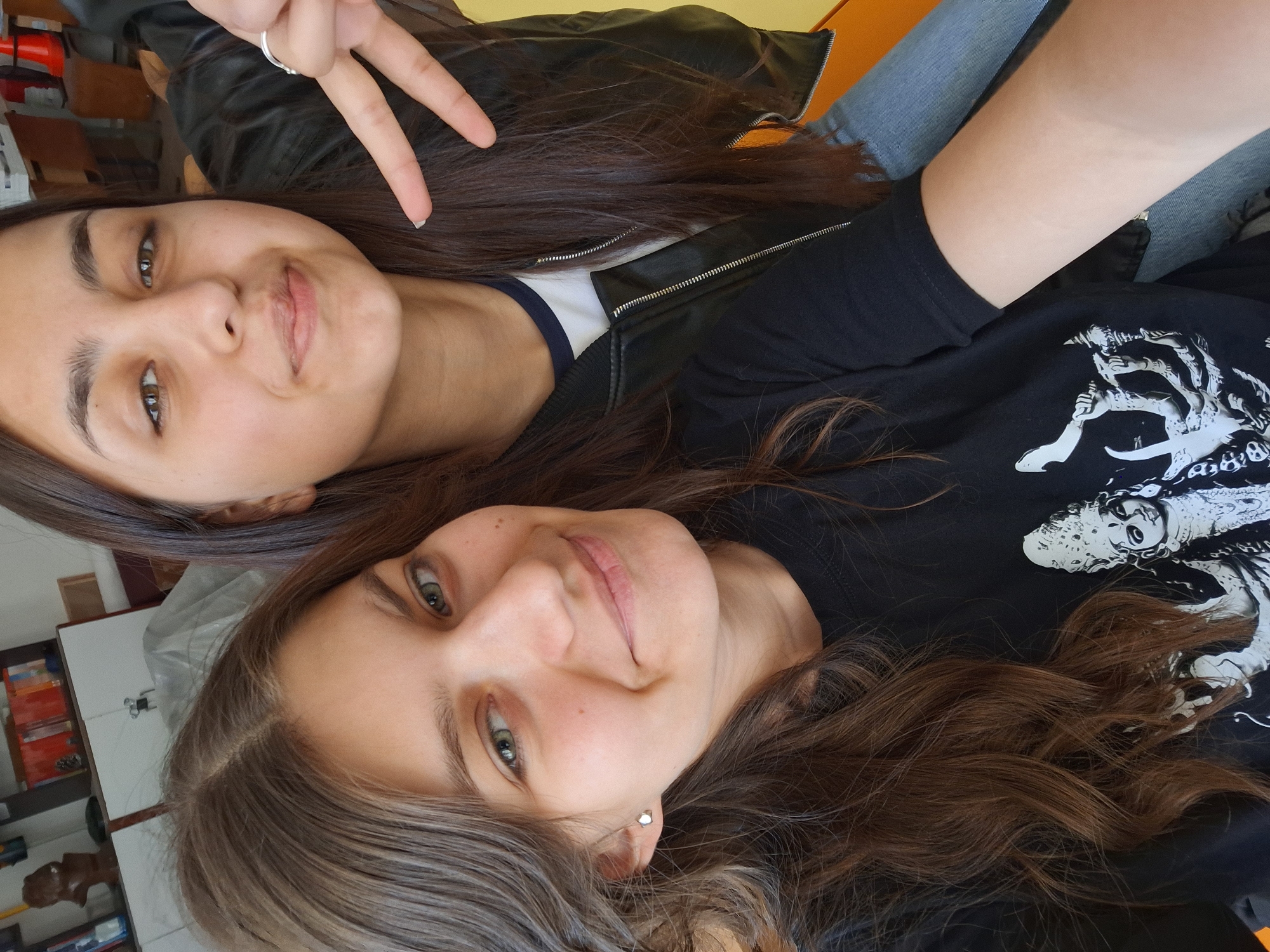 Two young women are posing closely together, seemingly taking a selfie. The woman on top is making a peace sign with her fingers, and she is wearing a black leather jacket over a white shirt with blue trim. She has brown hair and is wearing a ring on her finger. The woman beneath her is wearing a black t-shirt with a white graphic design on it. Both have brown hair and are smiling. The background is somewhat cluttered, with what appears to be a shelf with items and a hint of a window. There is also what appears to be furniture in the background. The location is Panagyurishte, Bulgaria.
