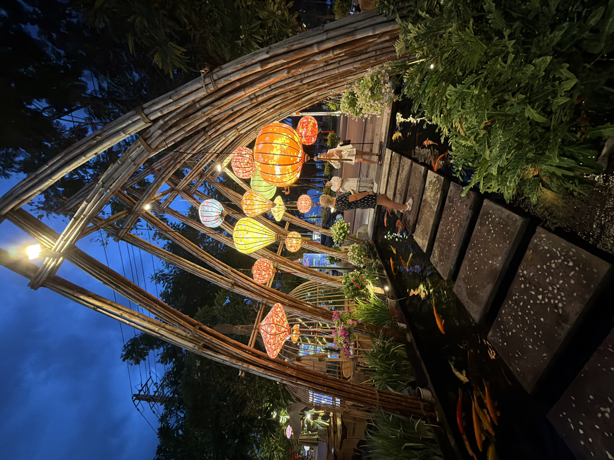 This image captures an evening scene in Nha Trang, Vietnam, likely at a restaurant or garden cafe. The foreground features a koi pond with several orange and white fish swimming. Stepping stones, made of dark rectangular slabs speckled with white, lead across the pond. Overhanging the pond is an archway constructed from bamboo poles, illuminated by colorful lanterns hanging from the structure. The lanterns are of various shapes and sizes, predominantly in red, orange, and yellow, casting a warm glow.
Two women are visible on the stepping stones. The woman in the foreground, wearing a black and white patterned dress and white sandals, stands facing away from the camera, looking towards the pond. Another woman, in a white dress, stands further back on the stones, holding a phone. Behind the bamboo archway, a street scene is visible with trees, utility poles, and what appears to be outdoor seating areas for a restaurant. The sky is a deep twilight blue, indicating the time is either dusk or early evening. The overall atmosphere is serene and decorative, suggesting a public space designed for relaxation and enjoyment.
