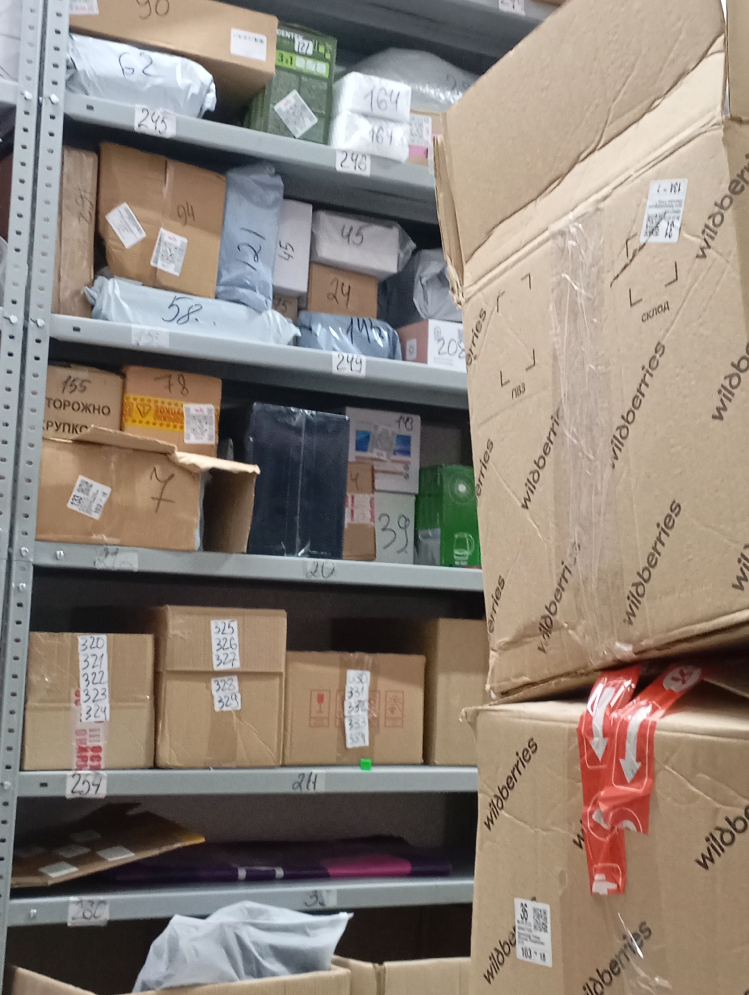 The image shows a warehouse or storage area, likely in Krasnodar, Russia based on the Wildberries branded boxes. Shelves are stacked with packages and boxes of various sizes and shapes, labeled with numbers. There is a visible metal shelf structure, with cardboard boxes bearing the "Wildberries" logo prominently displayed in the foreground. Other boxes are labeled with numbers such as "90", "62", "245", "94", "121", "45", "24", "143", "58", "208", "249", "155 TOPOЖHO", "78", "7", "20", "320-324", "325-329", "254", "214", "280" and "38". The overall environment is cluttered and organized for storage.