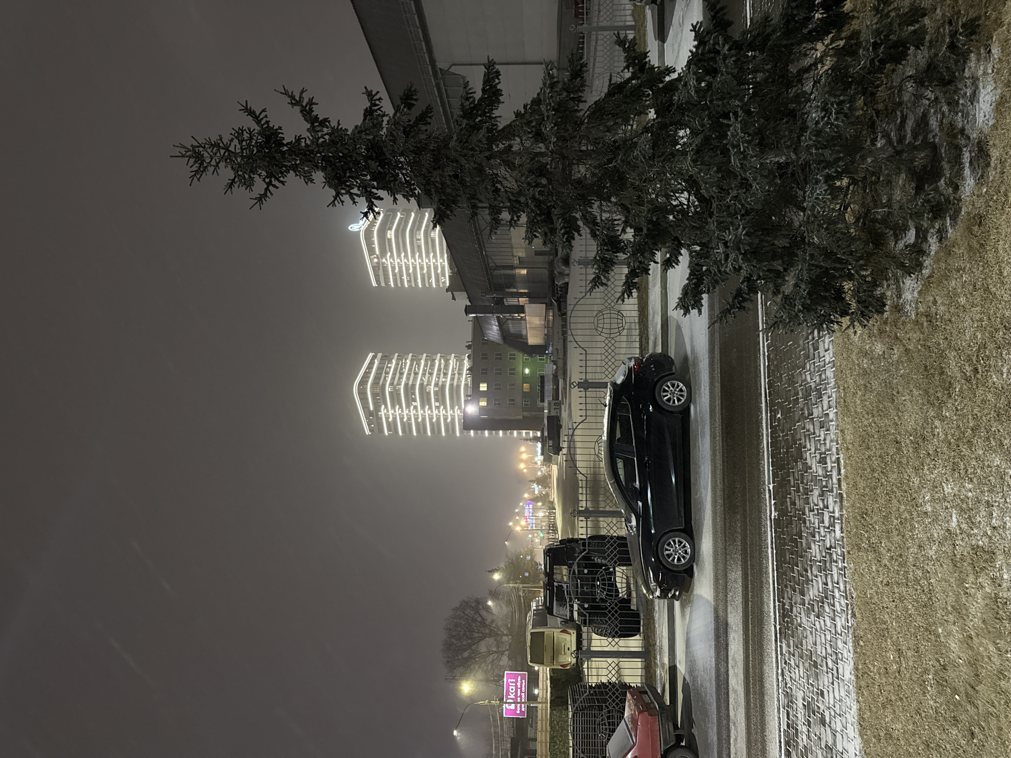 This is an exterior nighttime scene likely in the city of Krasnoyarsk, Russia, based on architectural style and signage. Two high-rise buildings in the background are illuminated with vertical rows of lights. A tree stands on the right side of the frame. A dark-colored car is parked on the street. There are other vehicles parked in a row, including a red car and a light-colored van. A sign with the word "kari" is visible. The environment appears to be snowy or icy, with evidence of snow on the ground.