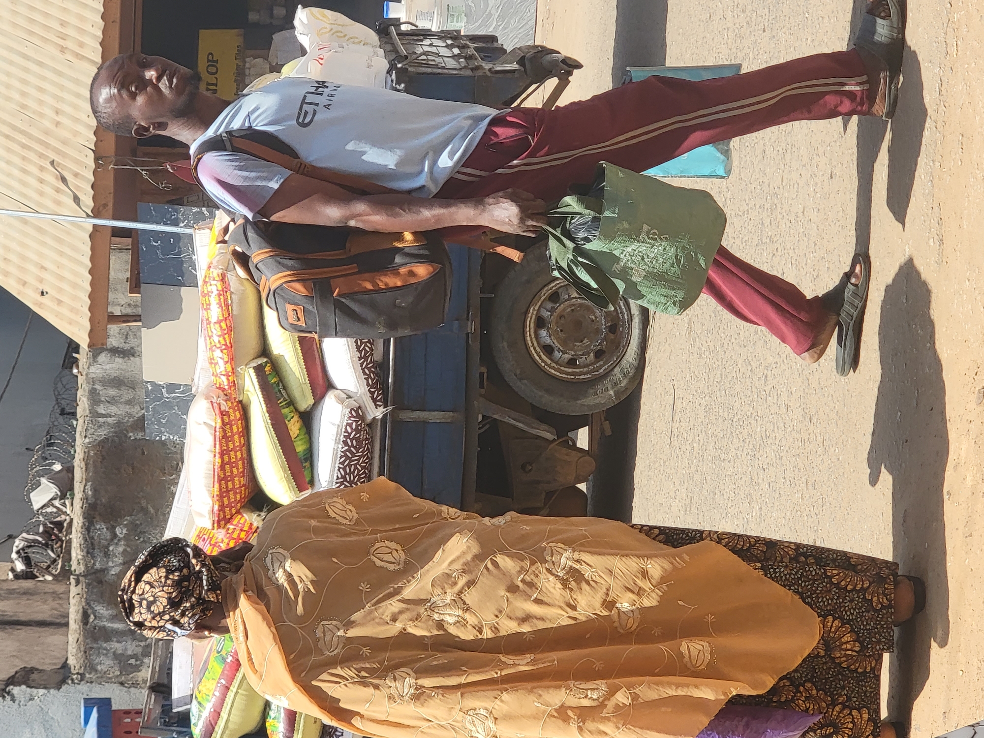 A man in a light blue t-shirt and red athletic pants stands beside a three-wheeled motor vehicle loaded with pillows, bags, and other items. He wears sandals and has a backpack on his shoulder. The vehicle is next to a woman wearing a gold-colored embroidered cloth, and it appears she is walking beside the vehicle. They are in a city with buildings and an asphalt road. Based on the prompt, the city is Maiburiji, Nigeria.
