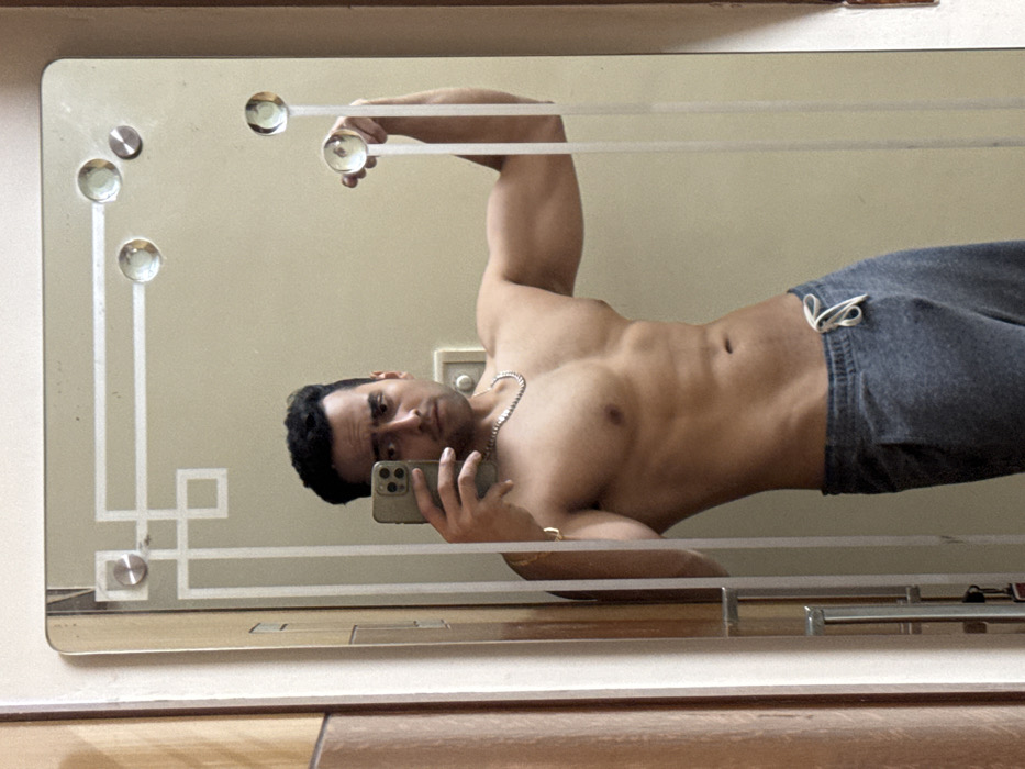 A muscular man poses in a mirror selfie, shirtless, with gray sweatpants. He holds an iPhone in his left hand. His right arm is raised, seemingly holding onto the top of the mirror. The mirror's design includes decorative glass orbs and etched lines. The background is a beige wall. He wears a chain necklace. The image is slightly rotated clockwise.