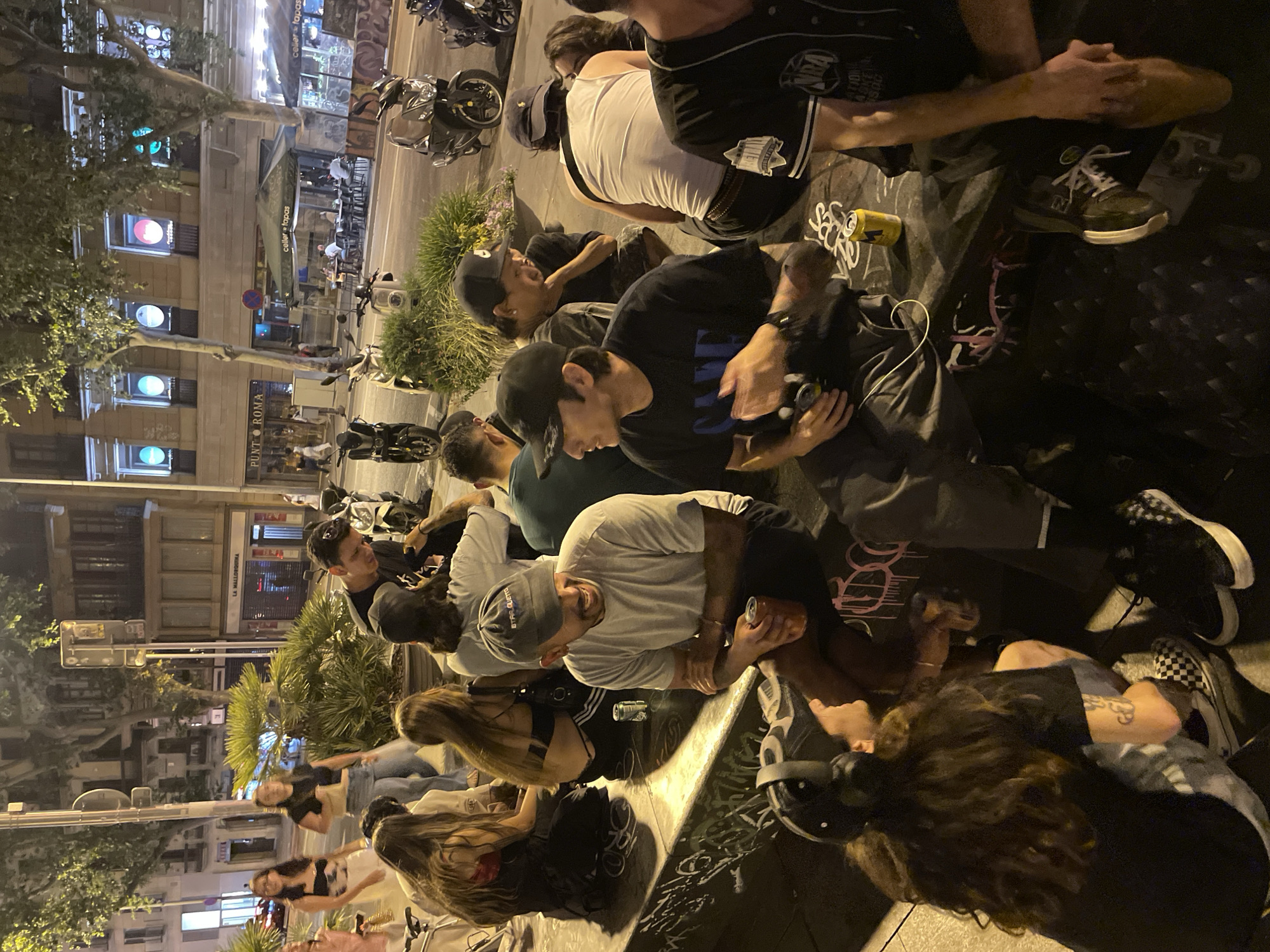 A group of people are gathered outdoors at night. Most of the people appear to be seated on the ground, close together. Many of them are young adults, and they are dressed casually. Several of them are holding cans or bottles.
The location is Barcelona, Spain, at the intersection of Carrer de Casp and Carrer de Girona. Streetlights illuminate the scene, and there are trees and buildings visible in the background. Several parked motorcycles are visible as well. Across the street is "Punt Roma" and "Celler + Tapas" is visible, indicating the presence of businesses in the area. Traffic lights are visible indicating the street's function as a thoroughfare.