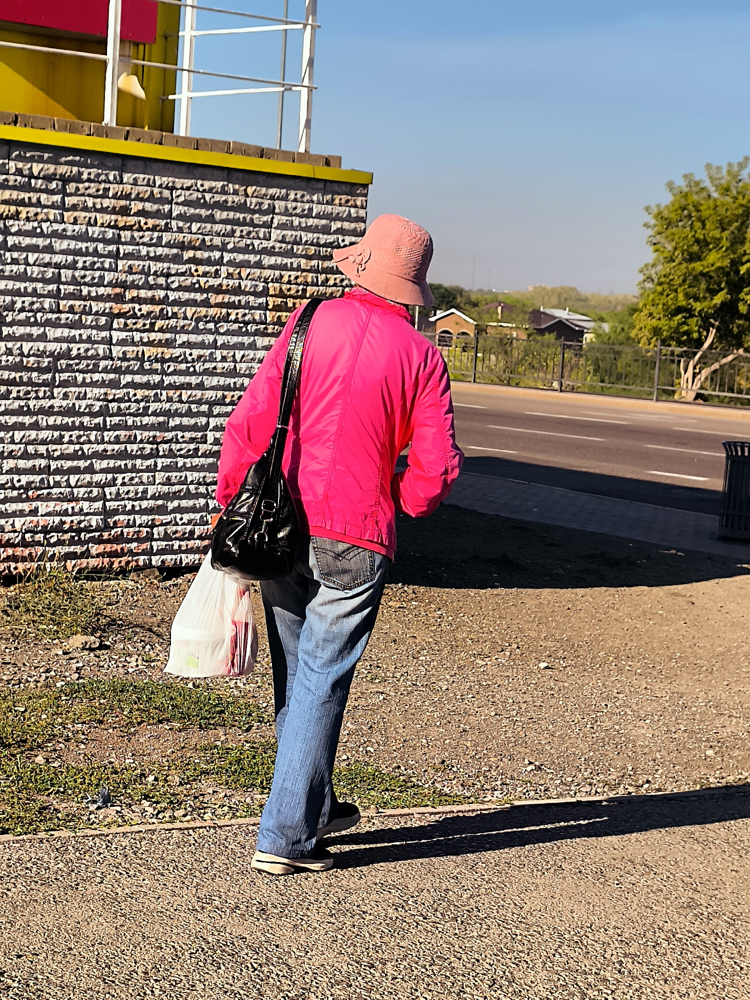 A person wearing a pink hat and a bright pink jacket walks on a gravel path beside a brick wall. They are carrying a black shoulder bag and a white plastic shopping bag. The person is wearing blue jeans and dark shoes. In the background, a road with white lines is visible, bordered by a fence and trees. The sky is clear and blue. There is no indication of the location being Qarağandı city administration, Kazakhstan.