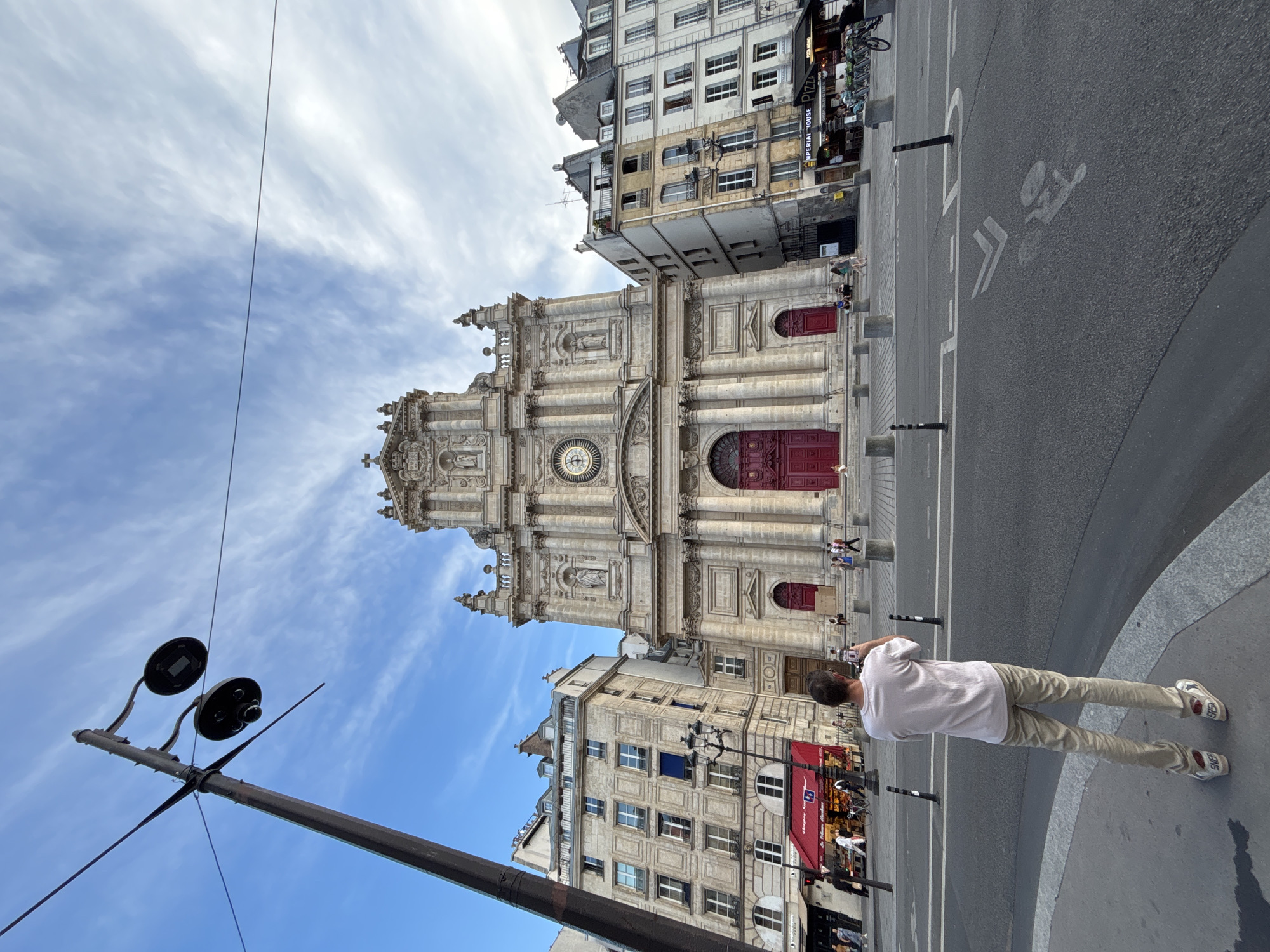An outdoor, eye-level, wide-angle shot captures the front facade of a stone church in Paris, France, under a partly cloudy blue sky. A man stands in the foreground, facing away from the camera, holding up a phone to take a photo of the church. He is dressed in a light pink t-shirt and tan pants, with white sneakers. The church features a large, ornate stone facade with a clock face at the top, a pediment, statues, and large red double doors at the entrance. To the left and right of the church are multi-story buildings with numerous windows. The street in front of the church is paved with asphalt and has white lane markings, including a bicycle symbol. Several streetlights are visible, including one to the left that appears to be holding two large lights and what might be a camera. People are walking and sitting on the steps of the church in the midground. There are also several shops with awnings and outdoor seating visible on the right side of the frame.