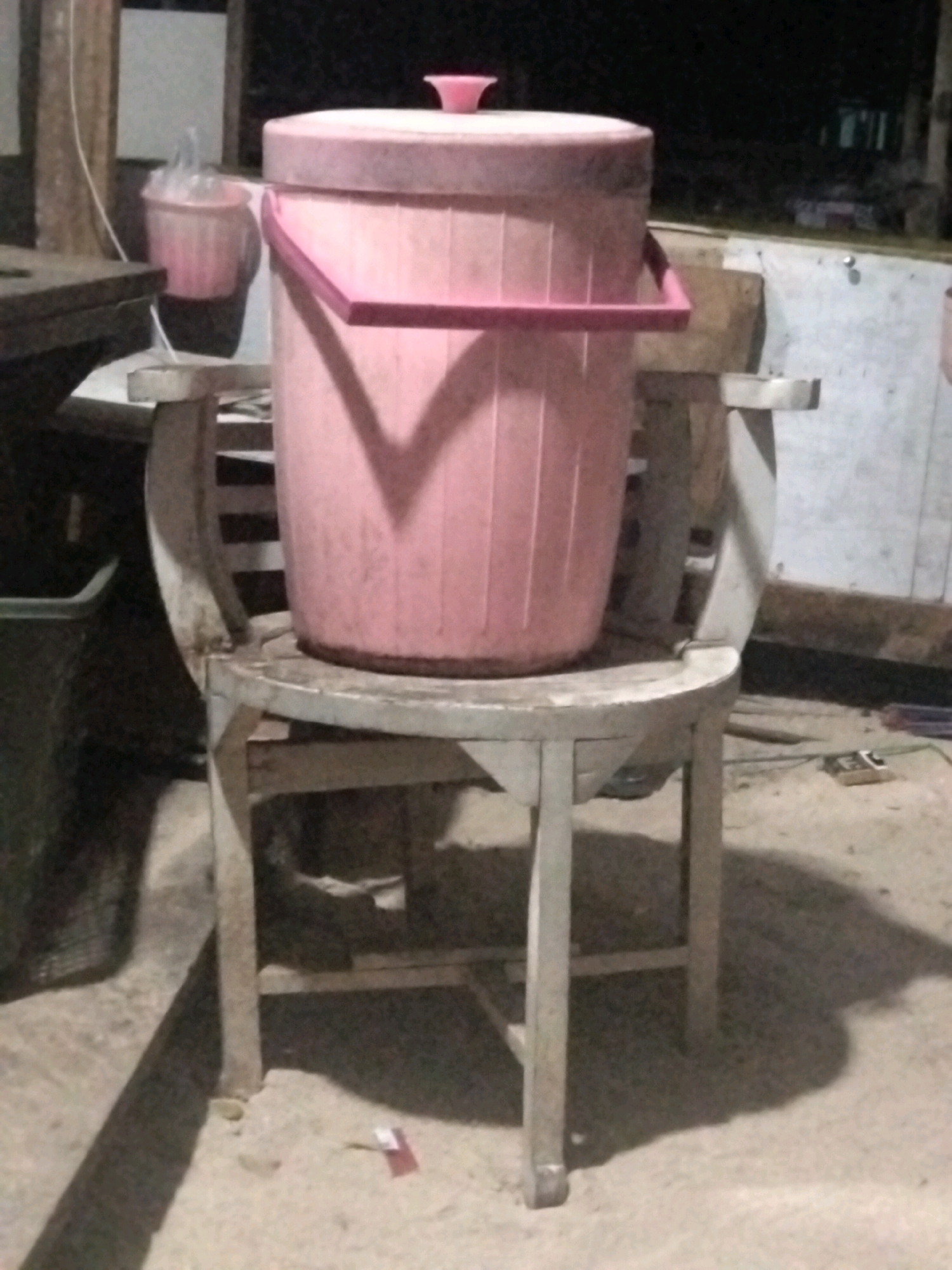 A pink plastic bucket with a pink lid rests on a wooden chair in an outdoor setting. The bucket is cylindrical with vertical ridges and has a pink handle attached to its sides. The chair is made of light-colored wood and has a rounded seat. The background includes a wooden structure, a wall, and some dark areas suggesting nighttime. A smaller pink bucket is visible to the left of the main subject. The ground is sandy and uneven, with shadows cast by the objects. The scene appears to be in Cikal, Indonesia.