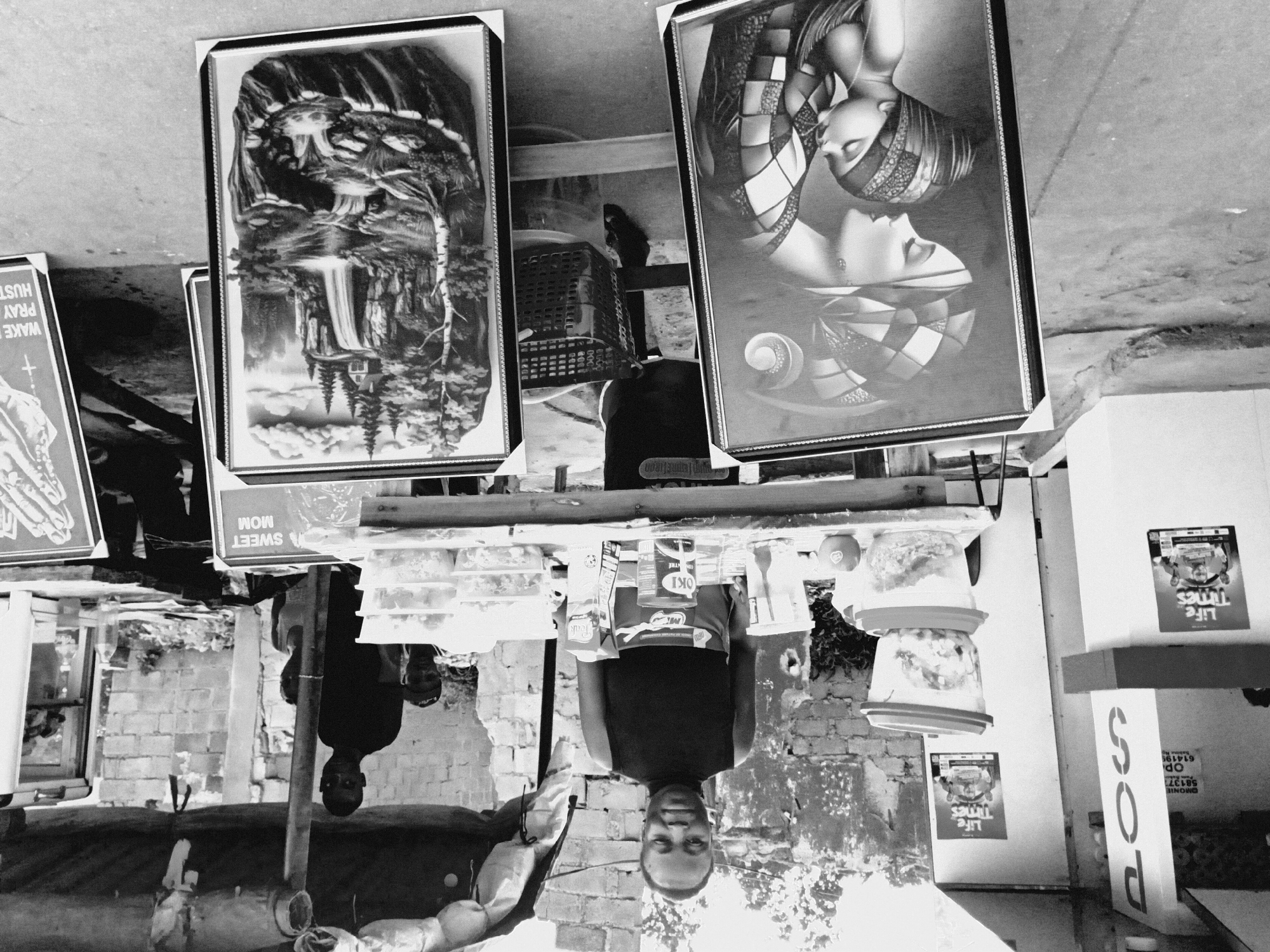 This black and white photo shows a street scene in Bukuru, Nigeria. Two framed pictures of artwork are visible on a table. One has a landscape with a waterfall, and the other contains an image of a woman. A man stands behind the table, with "OKI CENTRE" and "Teak" displayed on a product in front of him. Other items on the table include food and supplies. A sign above the table reads "A SWEET MOM," and nearby is another sign with the words "WAKE PRAY HUST." Posters reading "Life Times" are plastered on the wall of a building, where "POS" and a phone number, "581377," are painted.