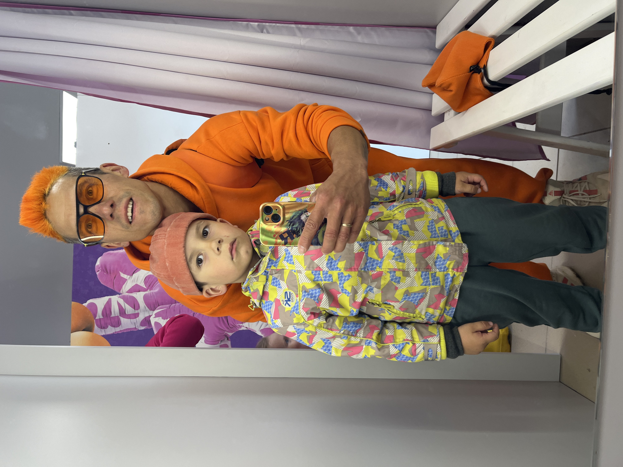 A man with orange hair and wearing an orange hoodie and sunglasses is taking a selfie in a dressing room with a child wearing a pink beanie and colorful jacket. The dressing room has a bench, a curtain, and a wall with a picture of people in ski gear. The image was taken in Chaikovsky, Russia.