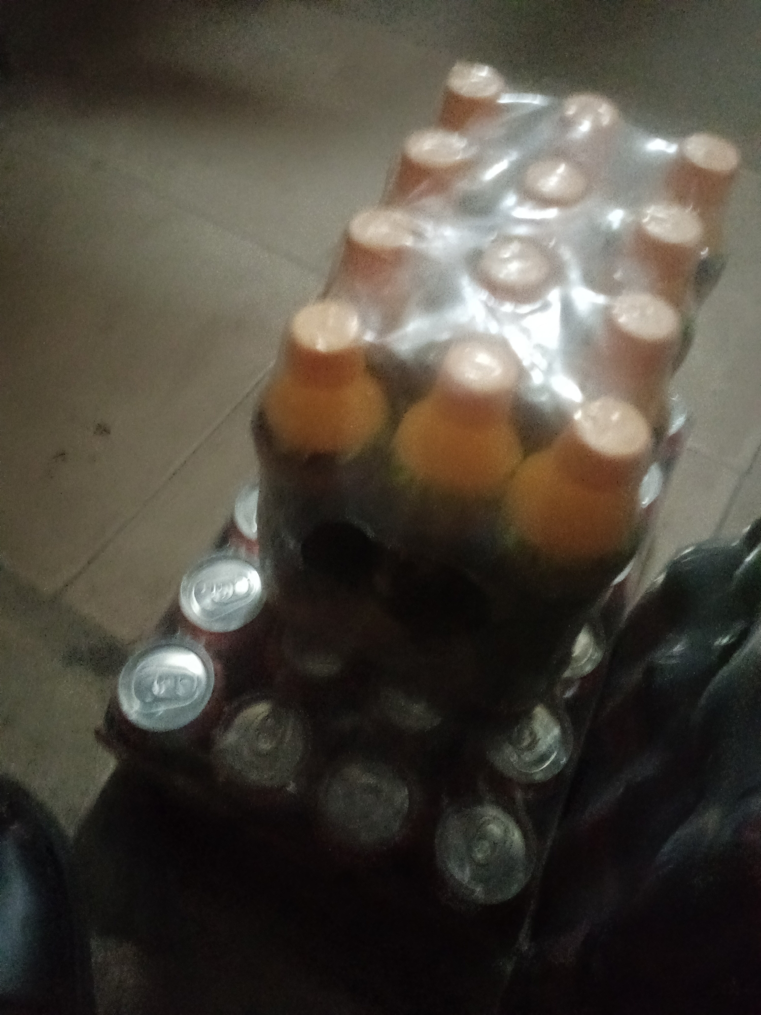 A group of bottled drinks, likely soda or juice, are shrink-wrapped together and placed on a tiled floor. The bottles have orange caps and dark liquid. Some cans of soda are also visible on the floor in front of the bottles. The scene appears to be indoors, possibly a storage area or a retail space. The lighting is dim, and the image is blurry, suggesting it was taken with a low-quality camera or in low light conditions. The location is identified as Jalingo, Nigeria.