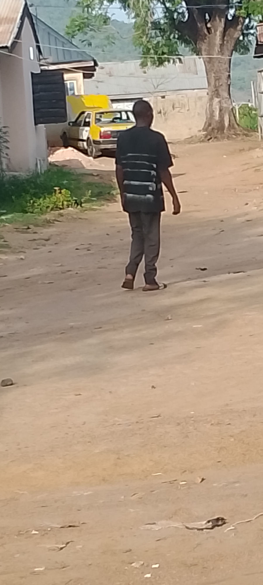 A person walks on a dirt road. The person wears a dark shirt with horizontal light stripes, gray pants, and sandals. Behind the person is a yellow car parked near a building. A tree stands to the right of the road. The location is Adankolo, Nigeria.