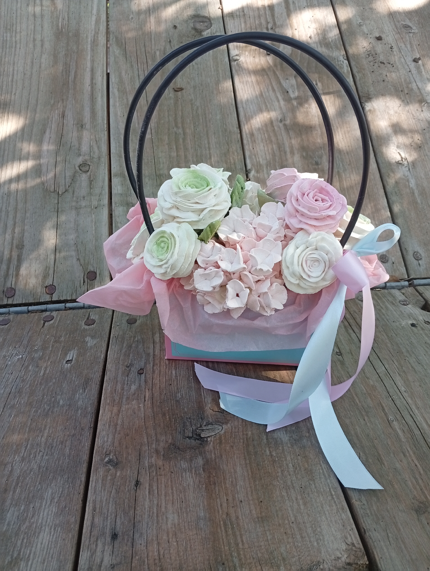 A floral arrangement in a box sits on a wooden surface. The arrangement consists of artificial flowers in shades of pink, white, and light green. Ribbons in light pink and white are tied to the arrangement. The wooden surface appears to be made of planks and has a rustic look with visible nails. The scene is lit by sunlight, casting shadows on the wood. The location is described as Unknown, Unknown.