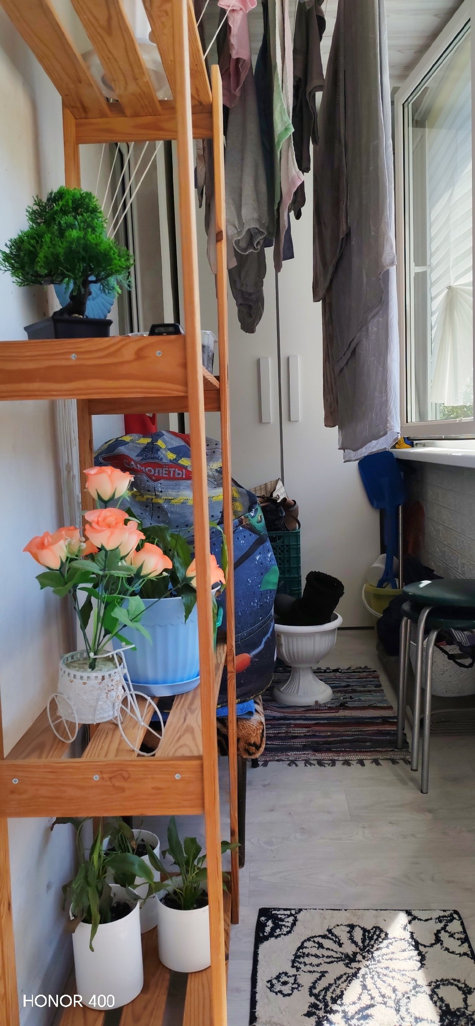 The image depicts a cluttered interior space, likely a balcony or storage room, in Noviy, Russia, as indicated by the text "САМОЛЁТЫ" ("Aircrafts") written in Russian on a bag. A wooden shelf is prominently featured on the left side, holding various plants in pots, including a small tree in a blue pot on the top shelf, roses in a blue pot on the second shelf, and three white pots with green plants on the bottom shelf. Behind the shelf, a partially visible wardrobe and hanging clothes create a sense of disorganization. A small pedestal with a pair of black boots, a rug, and a stool are positioned on the right side of the image. The floor is light-colored. A window with a partially drawn white curtain is visible in the background, providing natural light.
