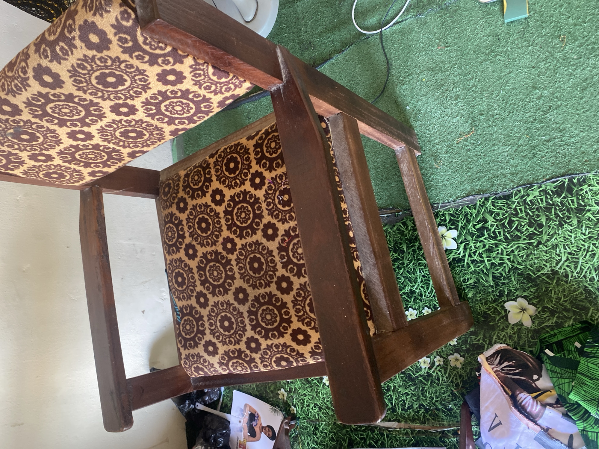 The image shows a wooden chair with a brown and tan patterned fabric upholstery. The chair has a simple, sturdy design with visible wooden joints. It is placed on a surface that appears to be artificial grass, possibly an outdoor rug or a decorative mat. Several small white flowers are scattered on the faux grass, adding a touch of nature to the scene. In the background, a white wall is visible on the left, and more artificial grass with a dark border can be seen on the right. A black plastic bag and some paper items, including what looks like a magazine or brochure with a person's image on it, are on the floor near the chair. Electrical cords are also visible in the background, suggesting an indoor or semi-outdoor setting. The lighting suggests it could be daytime. The overall impression is of a slightly cluttered but functional space, possibly a backyard or patio area in Jauro Yinu, Nigeria.