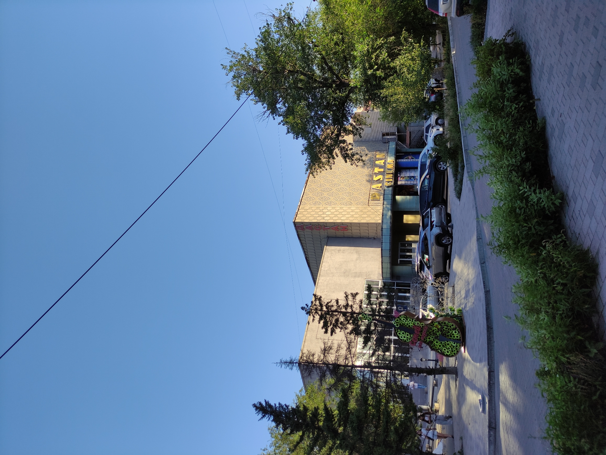 The image shows a frontal view of the "Astana Cinema" building in Semey, Kazakhstan, bathed in sunlight. The sky is clear blue, with a dark wire stretching diagonally across it. The cinema's facade is decorated with a patterned motif and large yellow lettering spelling out "ASTANA CINEMA." To the left of the entrance, there are several trees, including tall, slender coniferous trees and a larger deciduous tree to the right. In front of the cinema, a decorative sculpture resembling a large, green, floral-covered cello is visible, along with a couple of parked cars. Pedestrians are walking on the sidewalk in front of the building. The foreground features a paved walkway and a patch of lush green bushes. The overall scene depicts a daytime setting in a city environment.