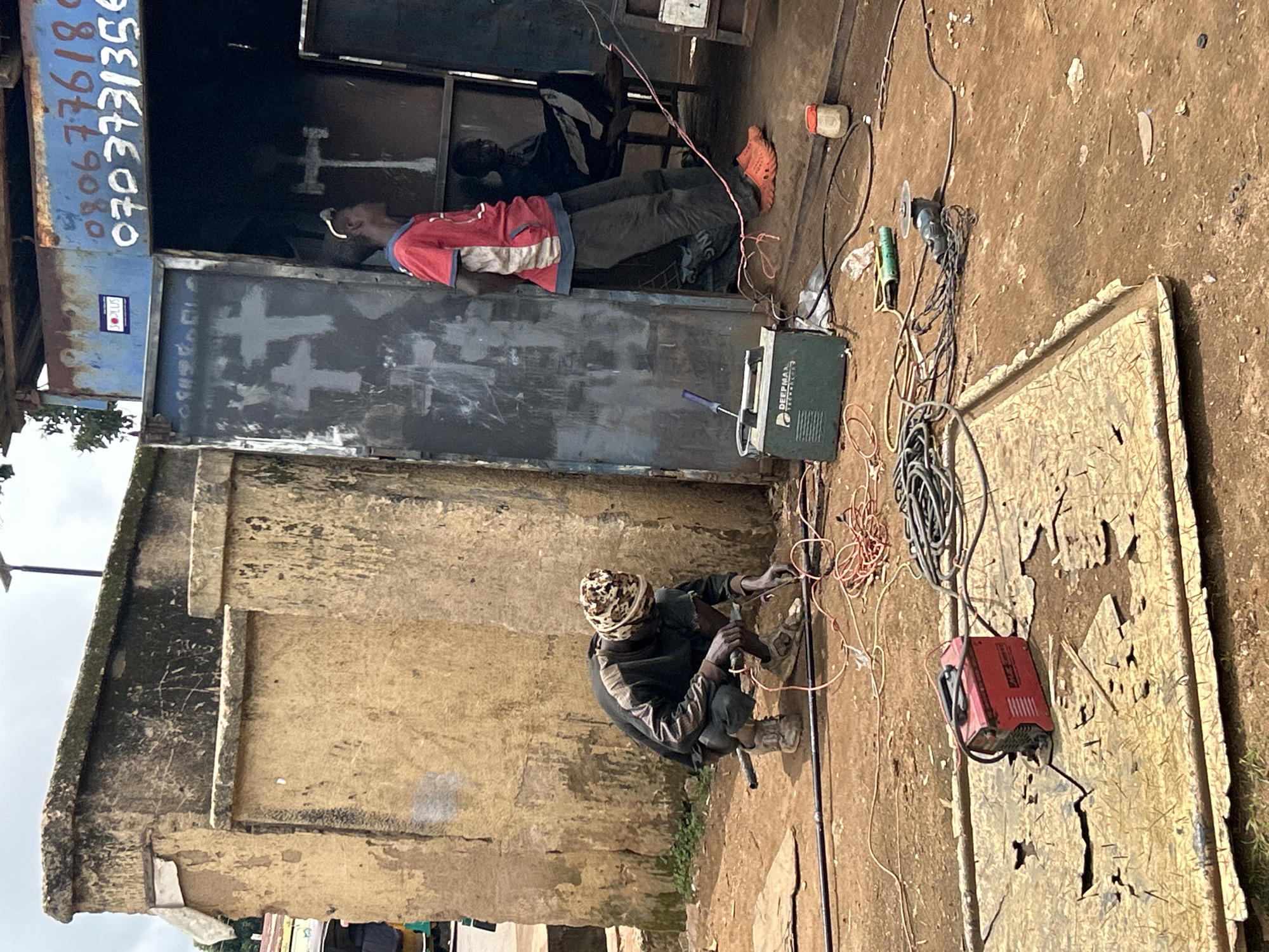 In Bar Jirgi Summa, Nigeria, an outdoor workshop scene unfolds. A man wearing a camouflage head covering, a dark long-sleeved shirt, and dark pants squats on the ground, appearing to be engaged in welding. He holds a tool in his right hand and has cables and a red welding machine nearby. To his right, a green welding machine is visible, along with various electrical cords and a small plastic container. In the background, a young man in a red t-shirt stands near an open doorway of a metal structure. Another person is seated inside the structure. The environment is dusty and appears to be a working area with scattered debris. The overall impression is one of a functional, albeit unrefined, industrial or repair setting.