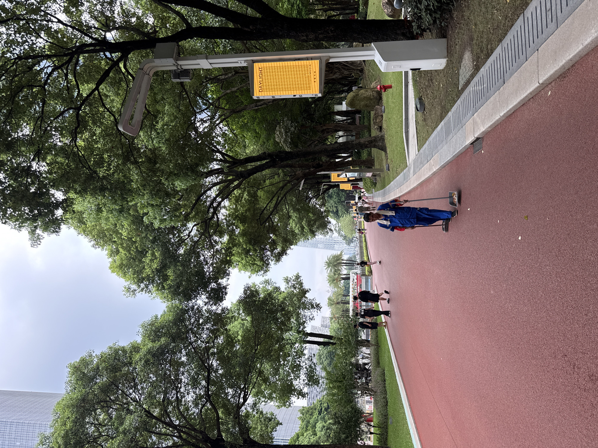 In a park setting in Tianhe District, China, a person in a blue uniform is pushing a cleaning cart along a red asphalt path. The path appears to be a running or cycling track, bordered by a grassy area and a concrete retaining wall with drainage slits. Mature trees with lush green foliage overhang the path, creating shade.
In the background, several other individuals are walking or running on the track. In the distance, modern high-rise buildings are visible, suggesting an urban environment. A yellow digital sign displaying "DAYLIGHT MEDIA" and some Chinese characters is mounted on a pole along the path. The sky appears overcast, indicating a cloudy day. The overall scene depicts a public park being maintained and utilized for recreation.