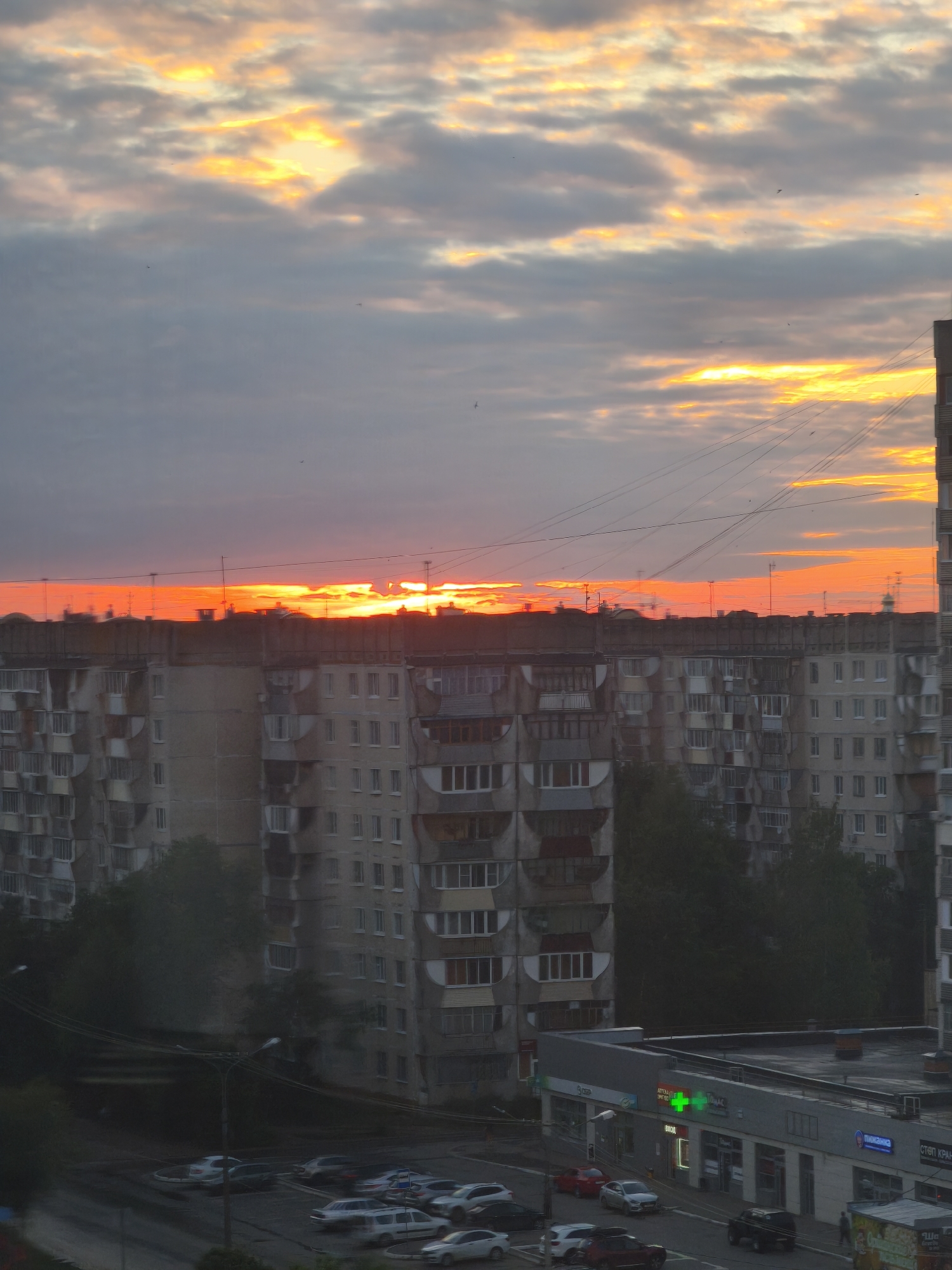 The image captures an urban scene in Yoshkar-Ola, Russia, during a sunset. The sky is filled with dramatic clouds in shades of orange, yellow, and gray, with the sun's rays breaking through. In the foreground, a row of multi-story apartment buildings dominates the landscape, their concrete facades showing signs of wear. Their balconies and windows are visible, some appearing dark and others reflecting the twilight sky.
Below the apartment buildings, a street is visible with several parked cars and a pharmacy with illuminated signage. The pharmacy has a green cross symbol and Russian text. Further down the street, more apartment buildings and trees can be seen. The overall impression is that of a residential area at dusk, with the vibrant sunset contrasting with the muted colors of the urban environment.