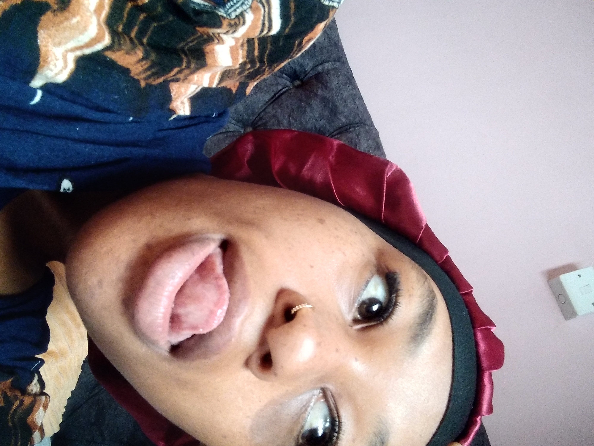 A young Black woman wearing a satin bonnet and a blue top with a patterned fabric overlay is captured in a close-up, selfie-style shot. She is looking away from the camera with her tongue extended. A diamond stud is visible in her left nostril. The background consists of a tufted grey couch and a pale pink wall with a white electrical outlet. The scene appears to be indoors, possibly in a residential setting. The woman's attire and the indoor environment suggest this image was taken in a private space, with no specific indicators pointing to the city of Nyeri, Kenya, or any other urban location.