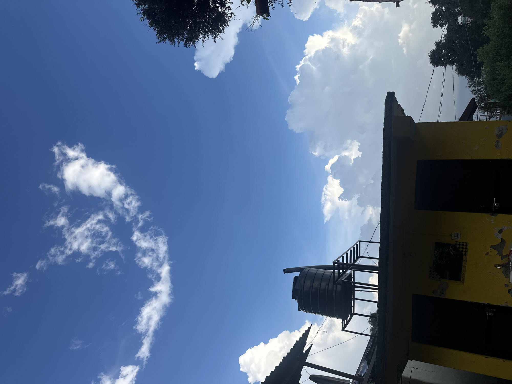 This is a low-angle, full-shot photograph of the sky and rooftops in Bhagsu, India. The sky is a vibrant blue, filled with a mix of fluffy white cumulus clouds and wispy cirrus clouds. The sun is not visible, but the clouds are brightly illuminated, suggesting daylight.
In the foreground, a portion of a yellow building with two dark doorways is visible. The paint on the building is peeling, revealing a darker layer underneath. Above the building, a large black water tank is mounted on a metal frame with steps leading up to it. The water tank appears to be made of plastic. To the left of the water tank, a corrugated metal roof is partially visible.
Further in the background, lush green trees and foliage can be seen, indicating a natural environment surrounding the buildings. There are also electrical wires strung between poles, typical of a residential area. The overall impression is one of a clear, cloudy day with elements of urban infrastructure integrated into a natural setting. There are no people or animals present in the image.