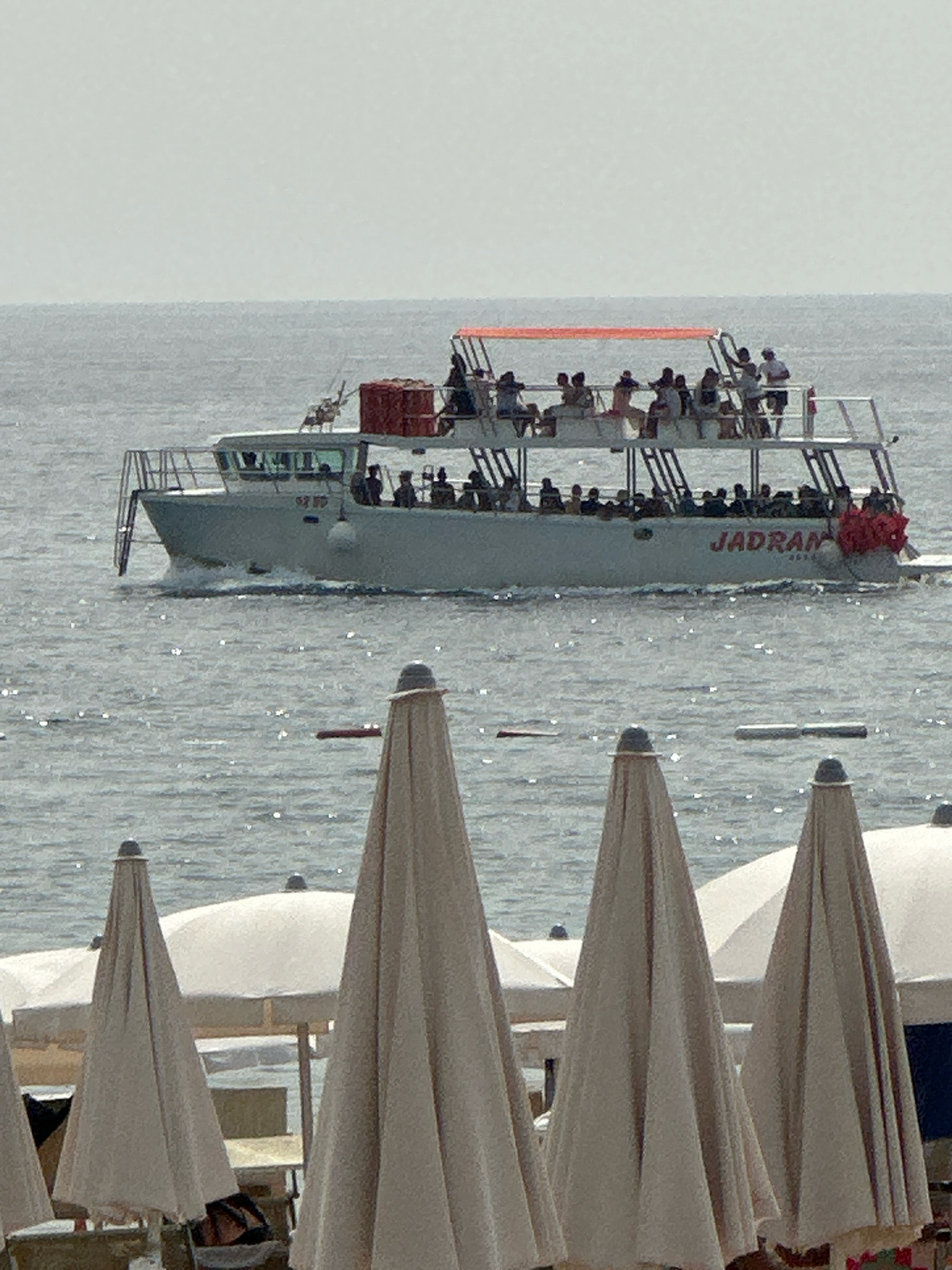 The image depicts a tourist boat named "JADRAN" sailing on a body of water, likely the Adriatic Sea, with the coastline of Boreti, Montenegro in the background. The boat is carrying numerous passengers on its upper deck and main deck. In the foreground, several closed beach umbrellas are visible, suggesting a resort area. The sky is overcast.