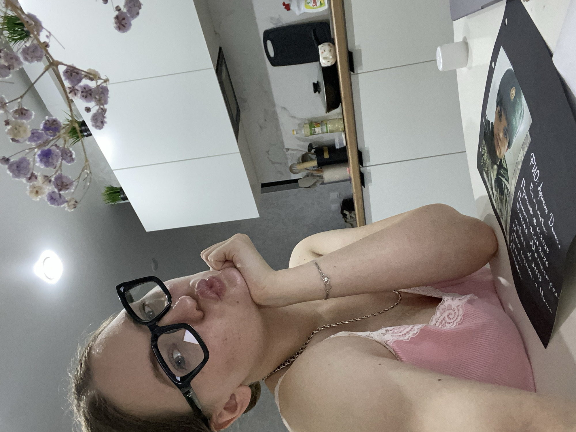 A young woman with glasses is pouting with her hand on her chin, looking at the camera. She is wearing a pink camisole with lace trim and a gold chain necklace. In front of her is a black folder with a printed photo and text. The background shows a modern kitchen with white cabinets and a marble backsplash. This scene is likely located in Izhevsk, Russia, as suggested by the presence of Cyrillic text on the folder.